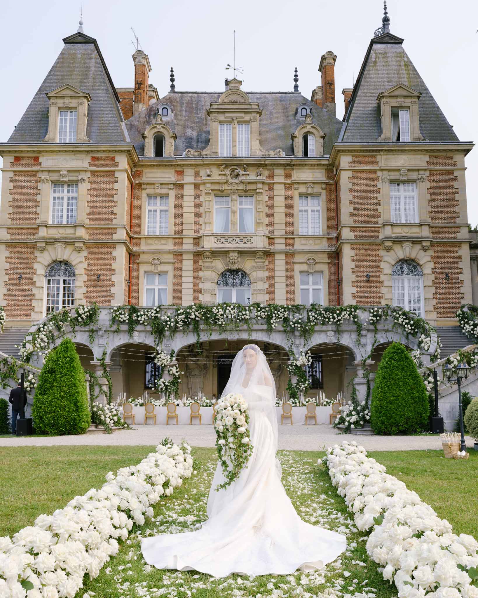 Bride in off-shoulder gown with cathedral train stands on lawn aisle lined with white peonies before French chateau