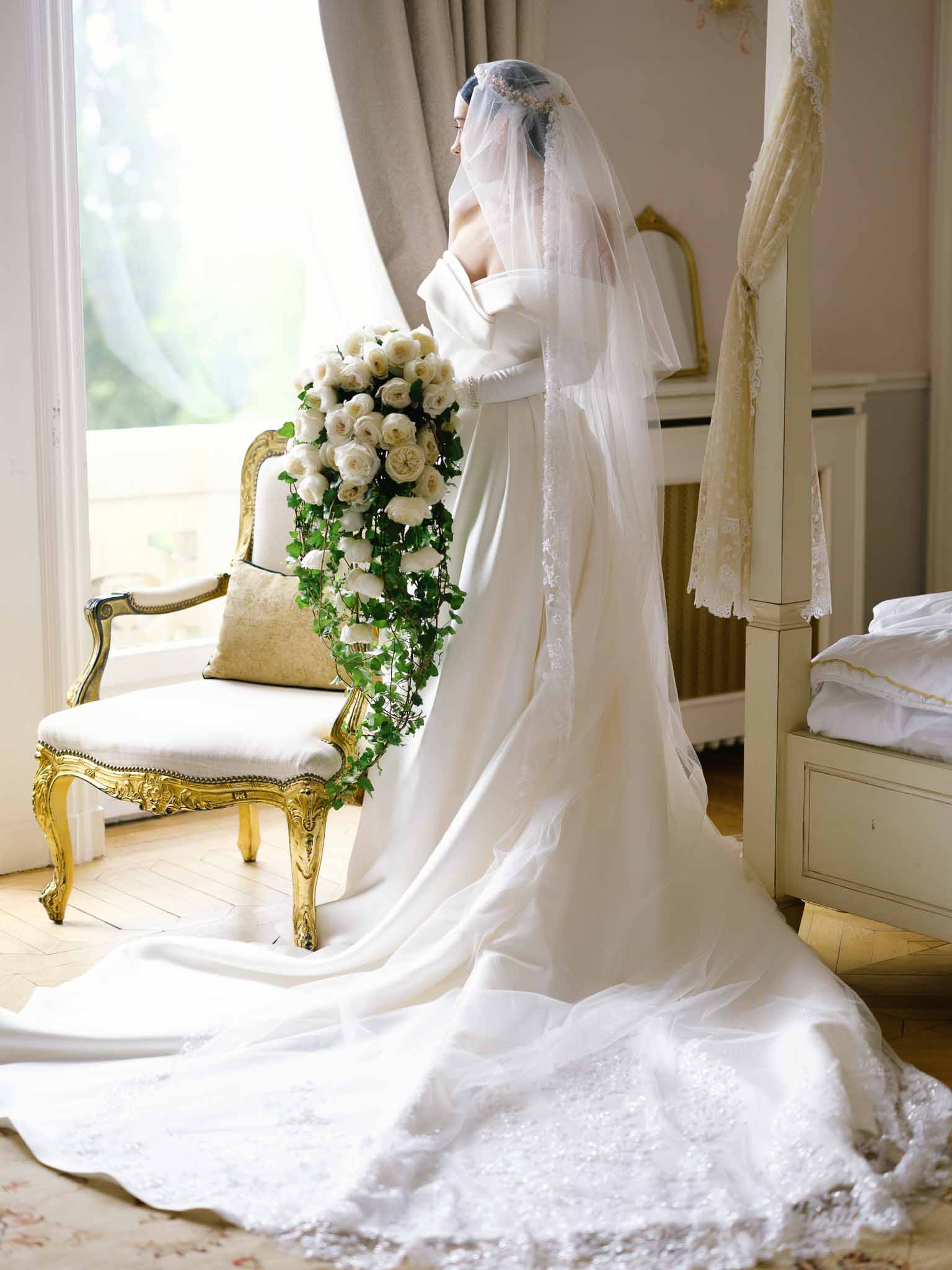 Bride photographed from behind near window wearing off-shoulder gown with cathedral train and veil holding cascading bouquet