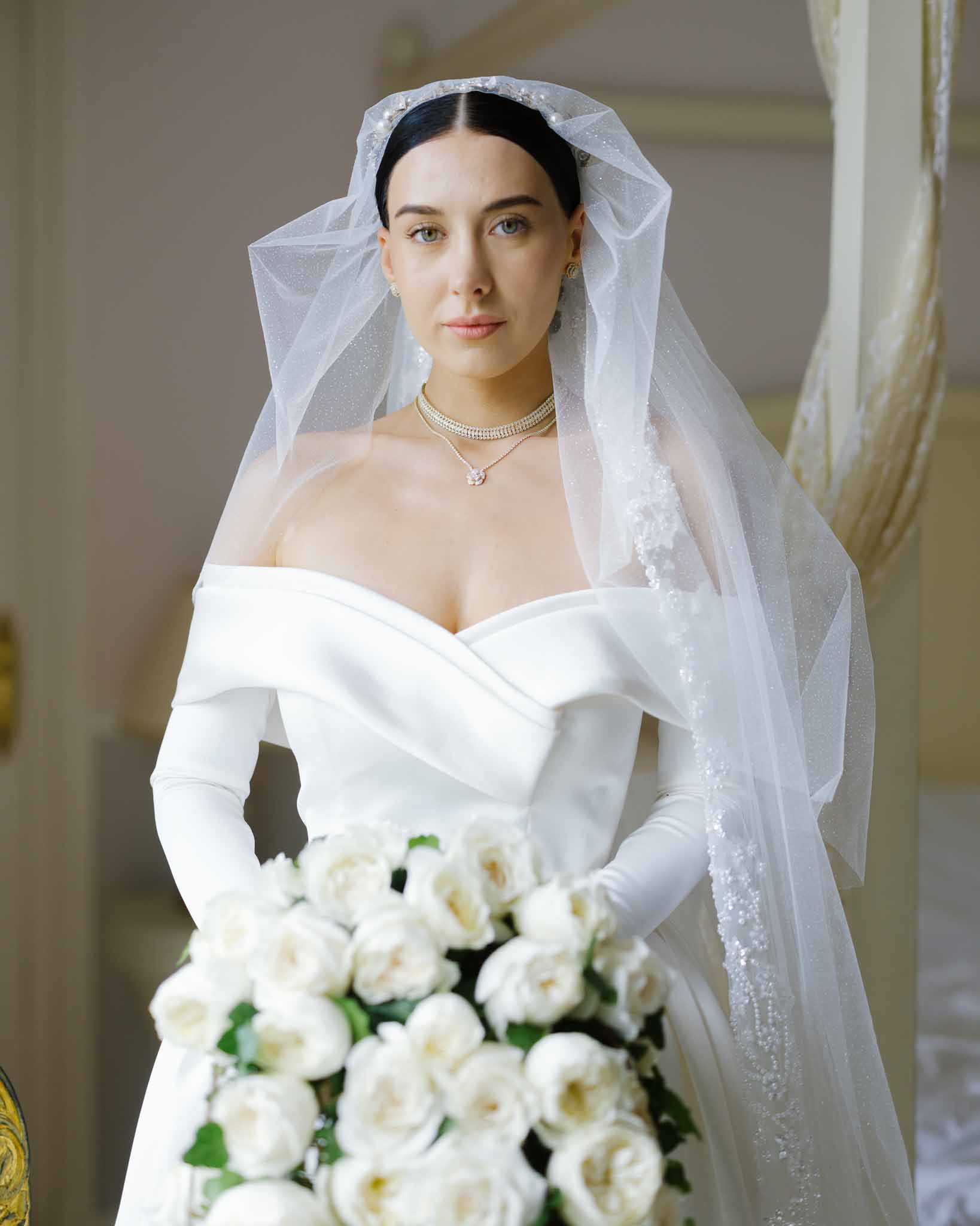 Bride in off-shoulder satin gown with crystal veil pearl choker and white garden rose bouquet indoors