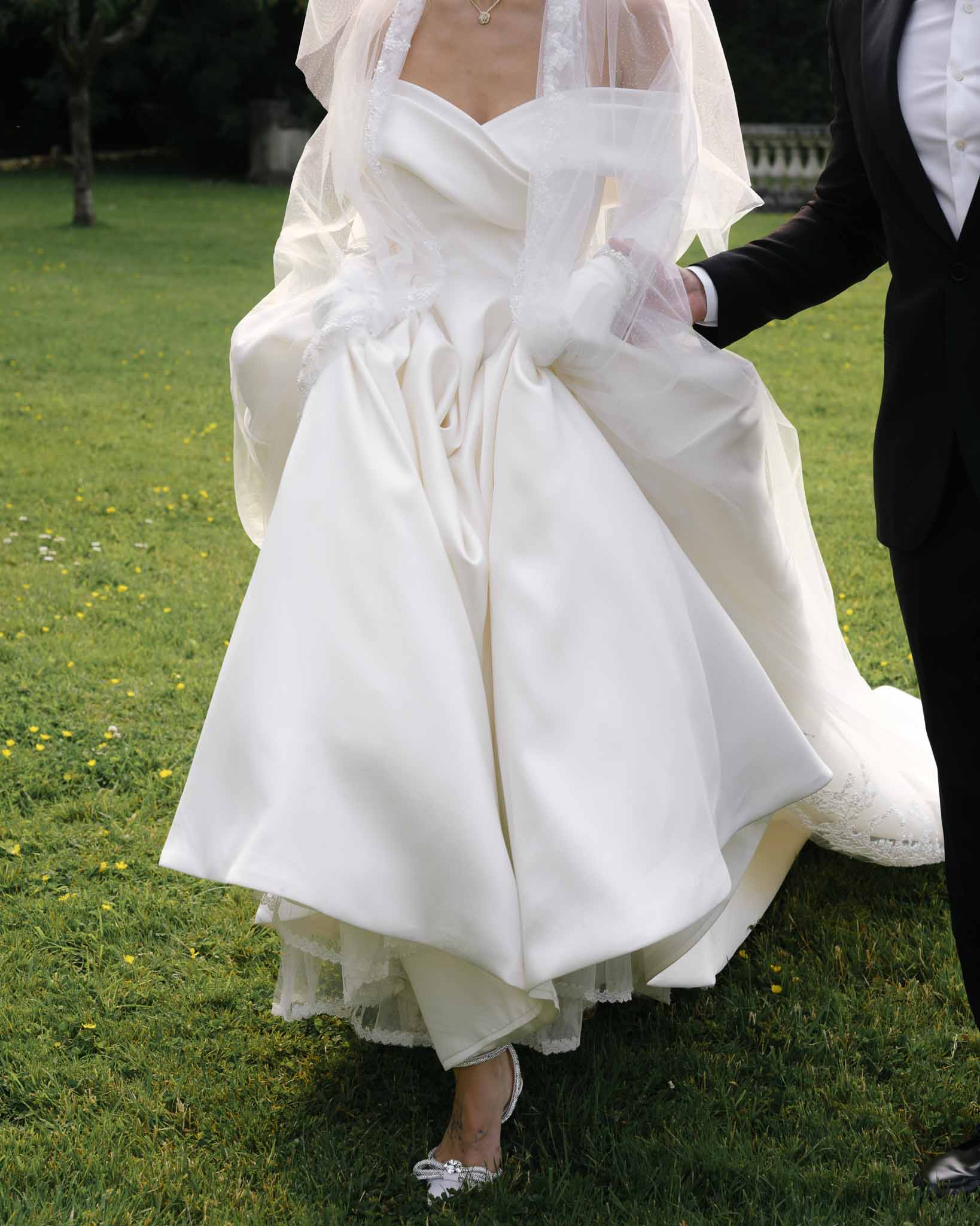 Cropped portrait of bride and groom walking on a garden lawn showing ivory satin ball gown, tulle gloves, and crystal heel...