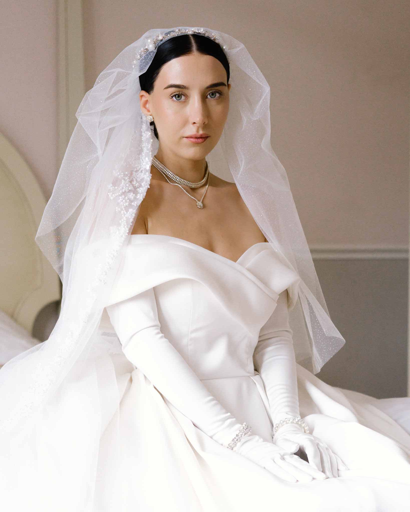 Seated bride in ivory off-the-shoulder satin ball gown with pearl headpiece, diamond choker, and tulle veil
