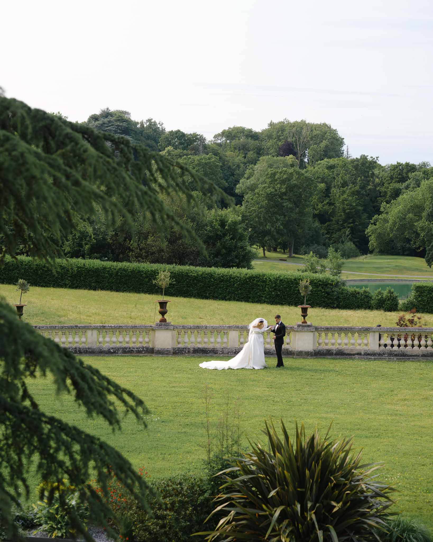 Bride and groom on chateau terrace grounds framed through cedar branches with cathedral train and veil visible