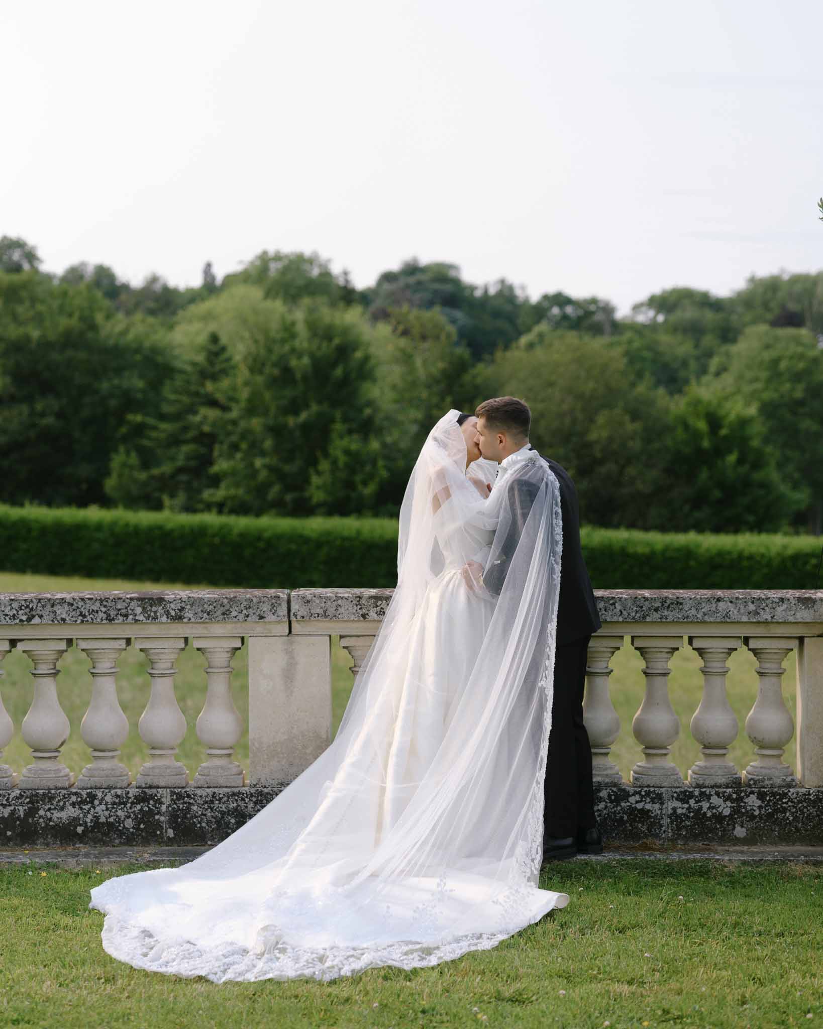 Bride and groom kissing on a stone balustrade terrace with cathedral-length lace train and veil from behind