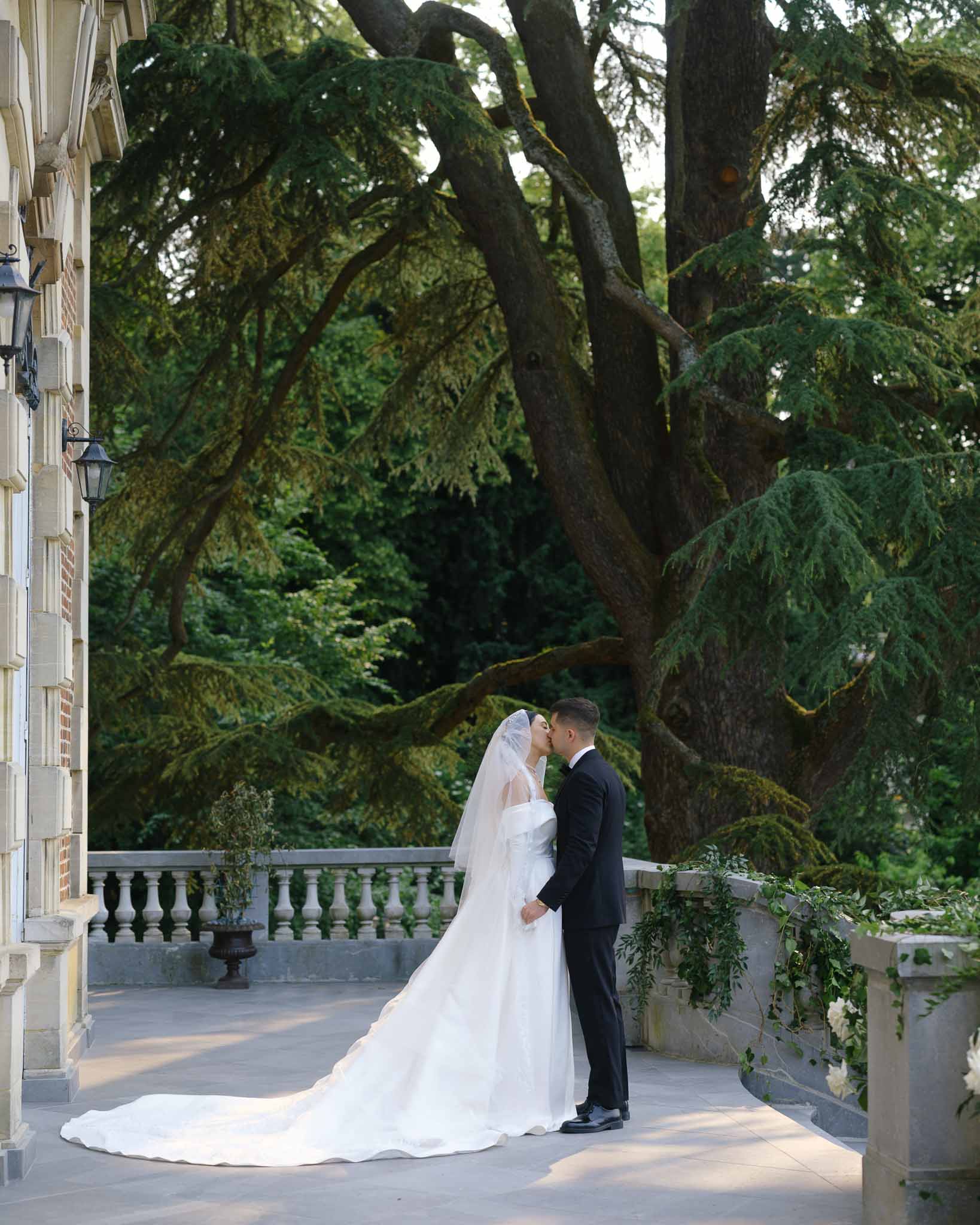 Couple kissing on stone balustrade terrace with cathedral train and lace-edged veil extending across paving
