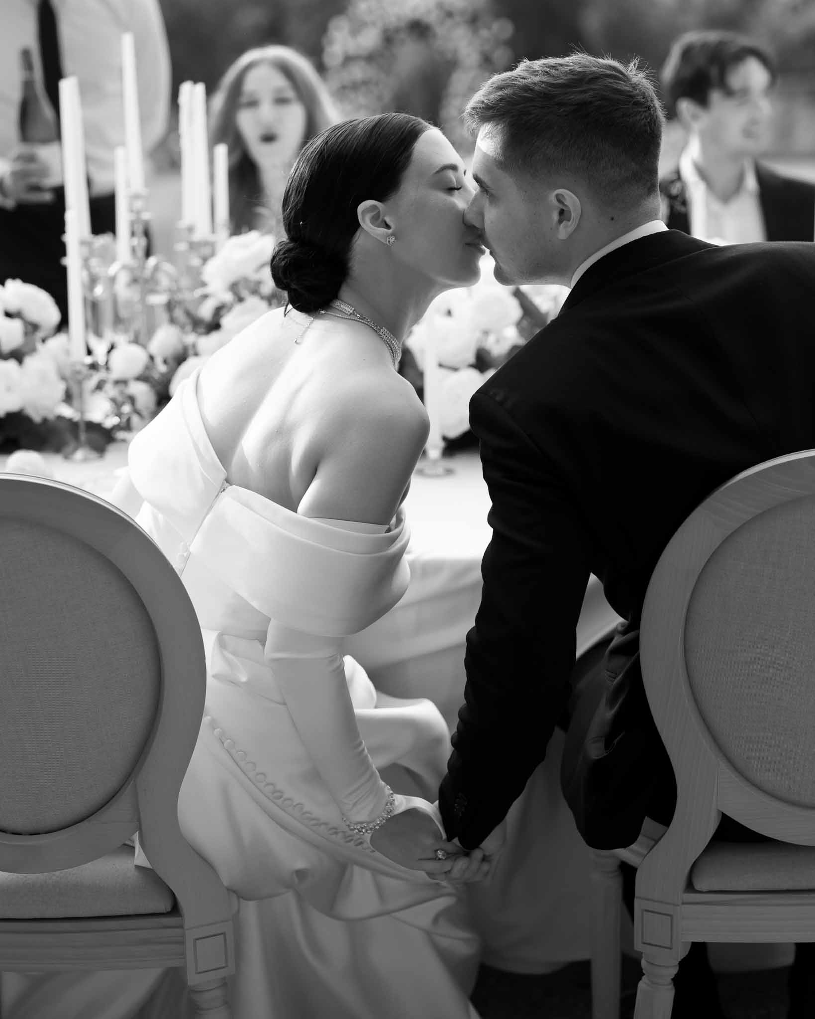 Black and white photo of bride and groom kissing at head table during wedding reception with candelabras