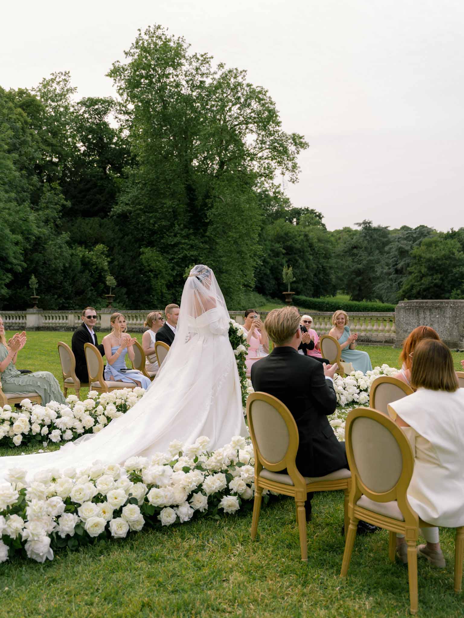 Bride walks down rose-lined grass aisle toward altar with guests in gold chairs at chateau garden