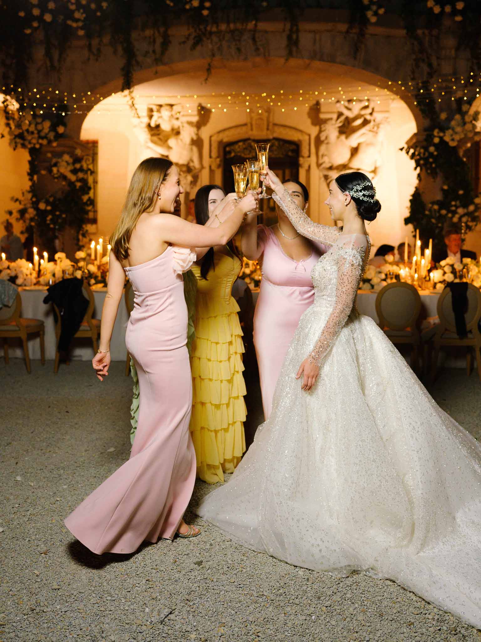 Bride in embellished ballgown toasting champagne with three guests during evening chateau courtyard reception