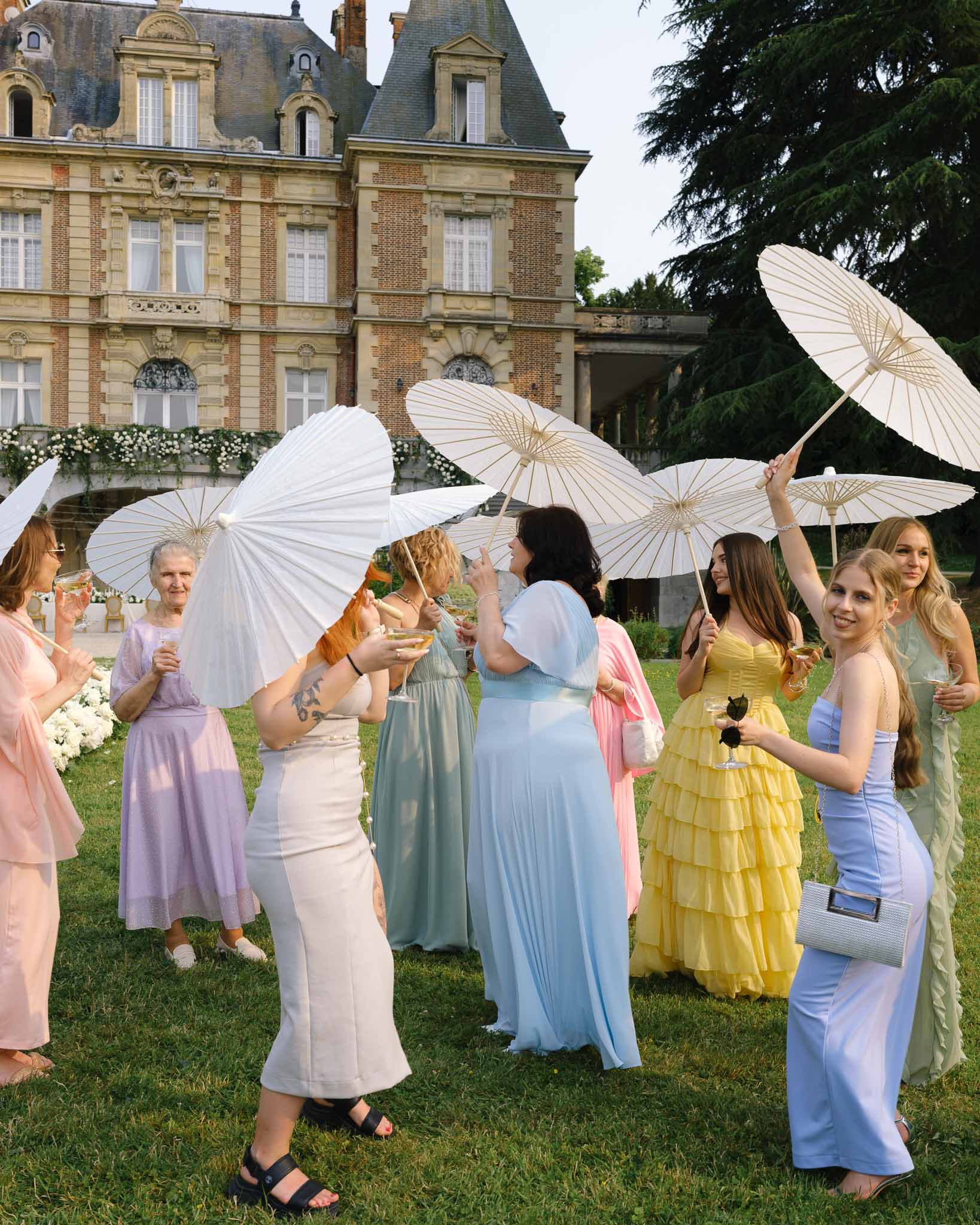 Ten female guests in pastel dresses holding parasols and champagne on chateau lawn