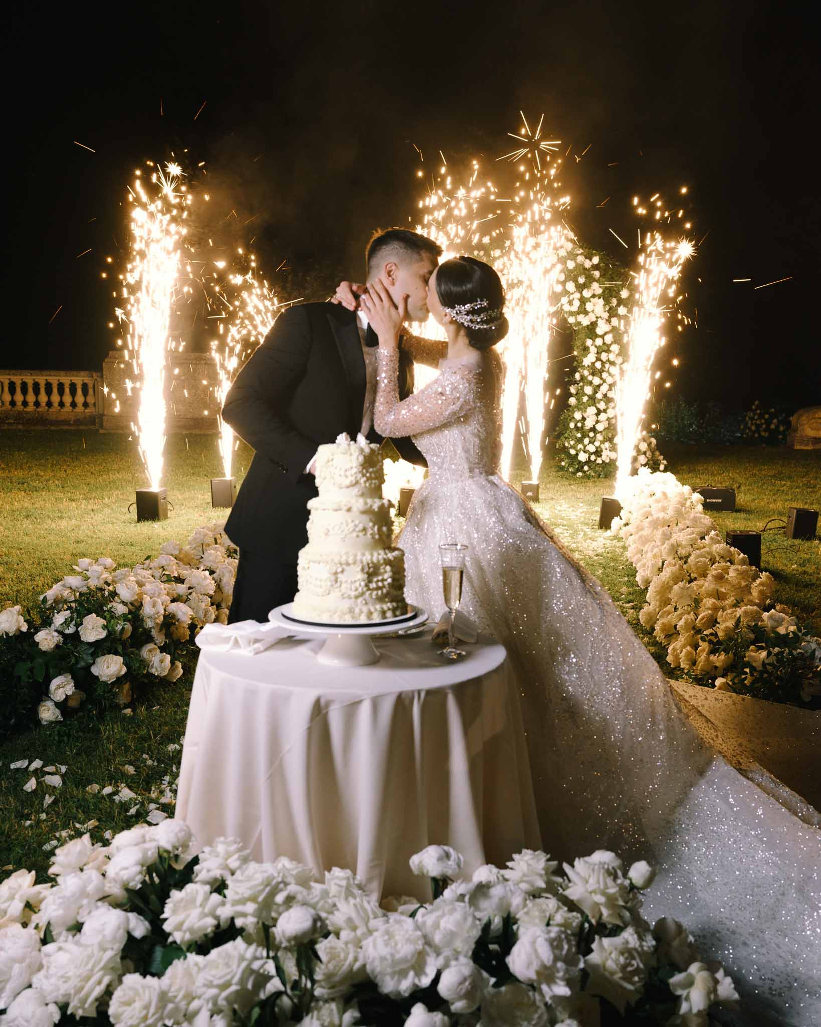 Couple kissing at four-tier pearl cake with cold spark fountains and white rose arch at night reception