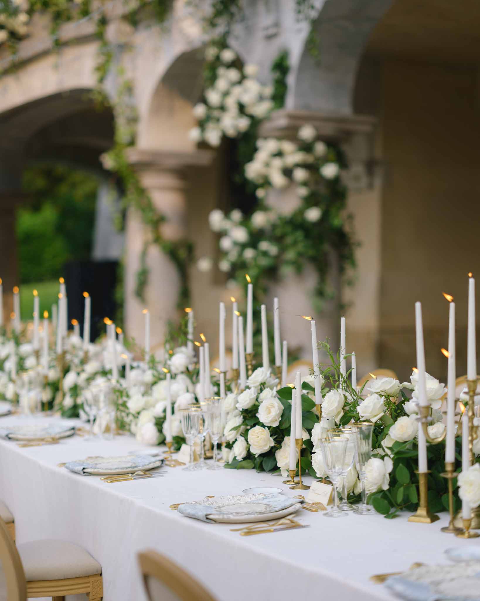 Reception table with white rose and eucalyptus runner, gold candlesticks beneath stone colonnades