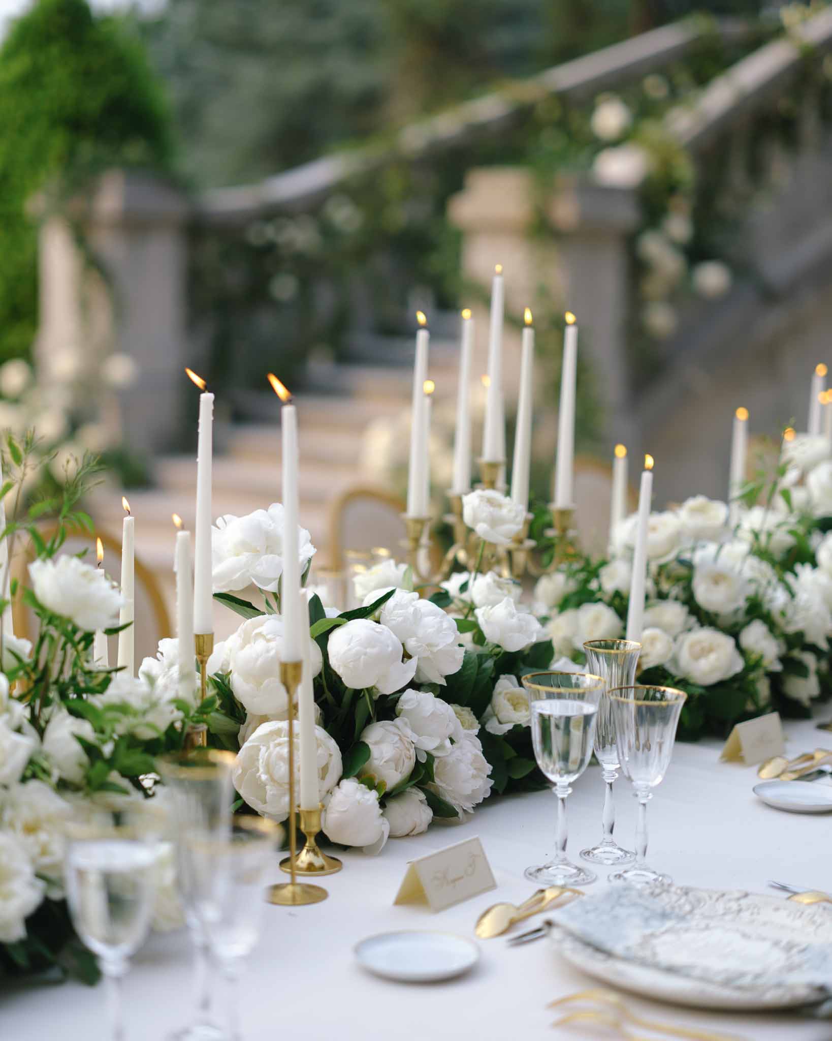 White peony runner with gold candelabras, gold flatware, and crystal glassware under greenery-draped pergola