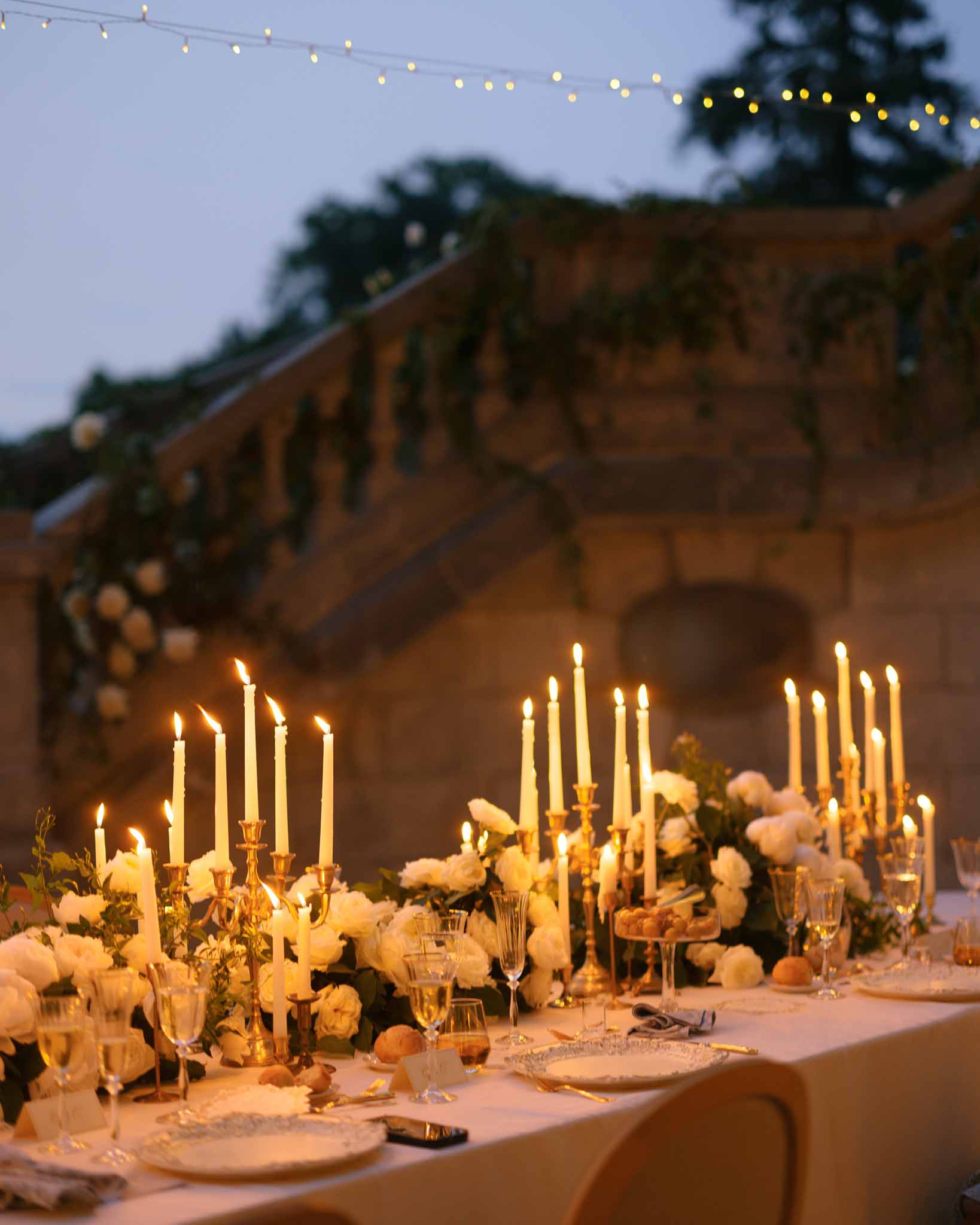 Evening reception table with gold candelabras, white roses, and fairy lights against stone staircase