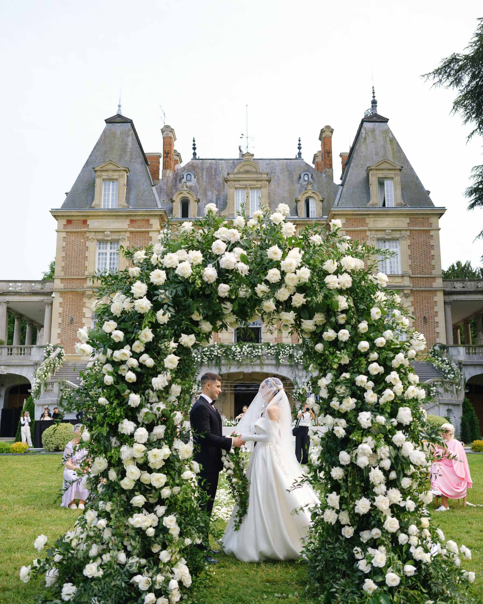 Couple exchanging vows under circular white rose arch on lawn before mansard-roofed chateau