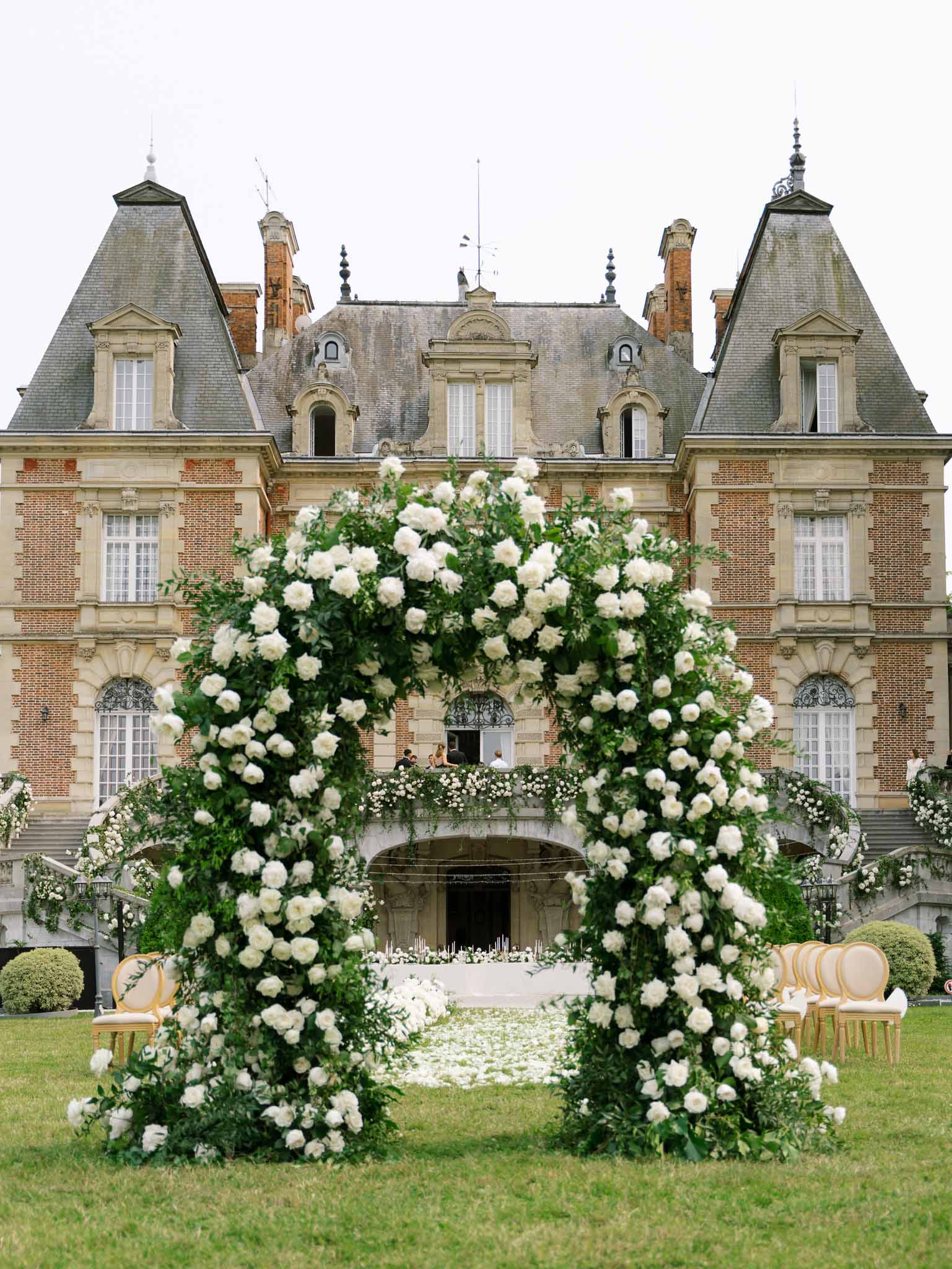Circular white rose arch with petalled aisle and black tapers before red-brick Renaissance chateau