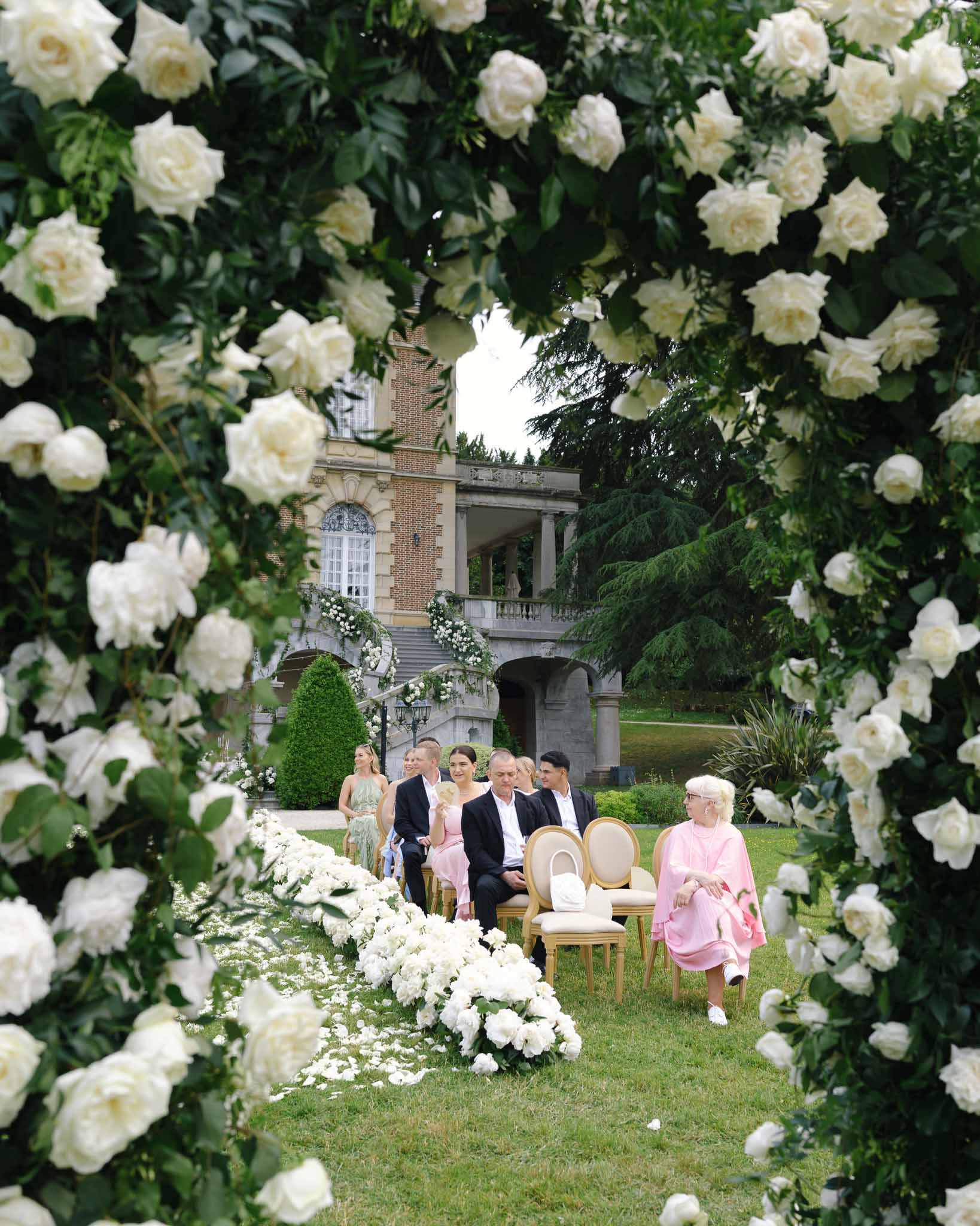 Ceremony through cream rose arch frame with gold chairs and white peony aisle before brick manor with balustrade