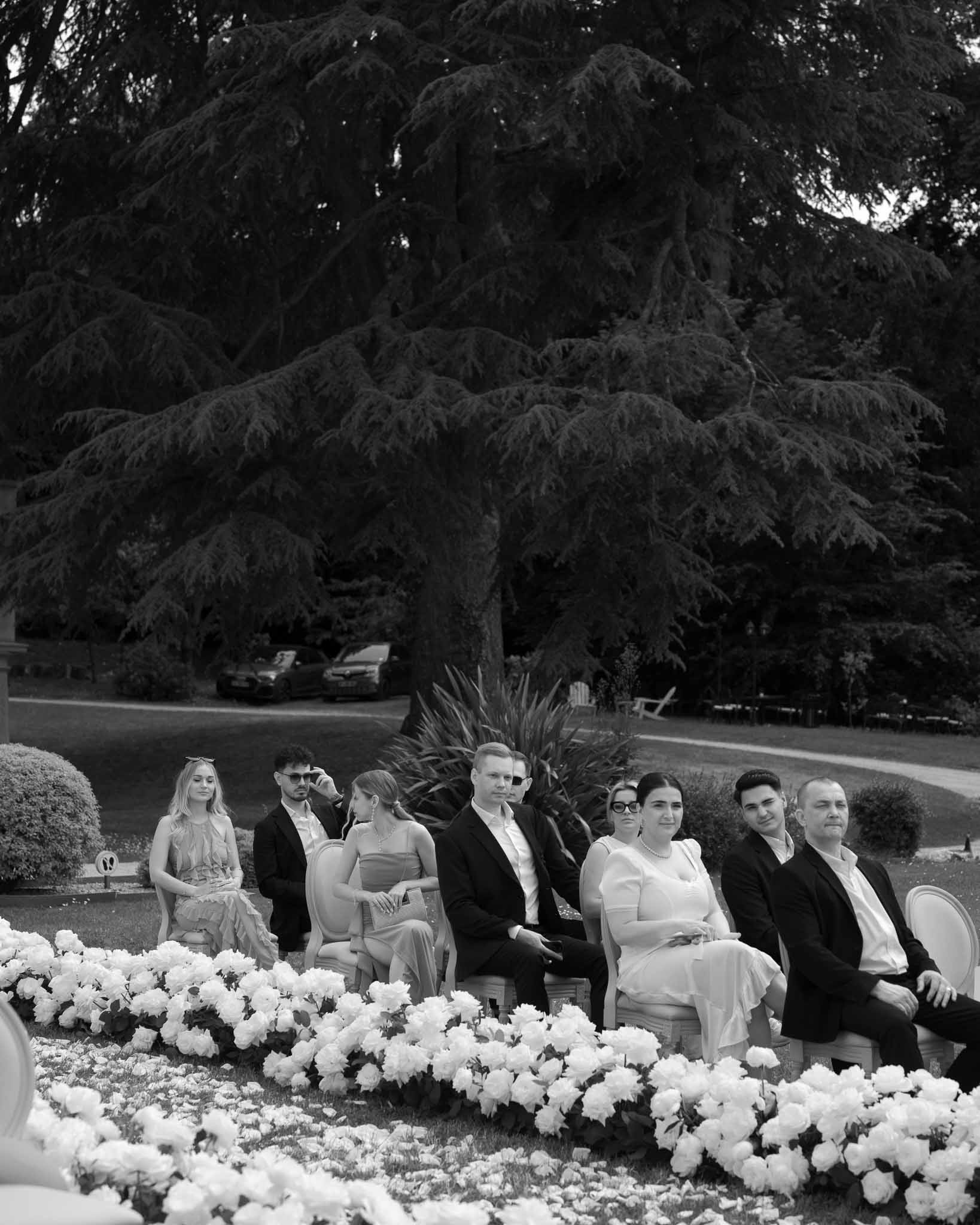 Black and white photo of wedding guests seated on lawn with lush floral aisle borders during outdoor ceremony