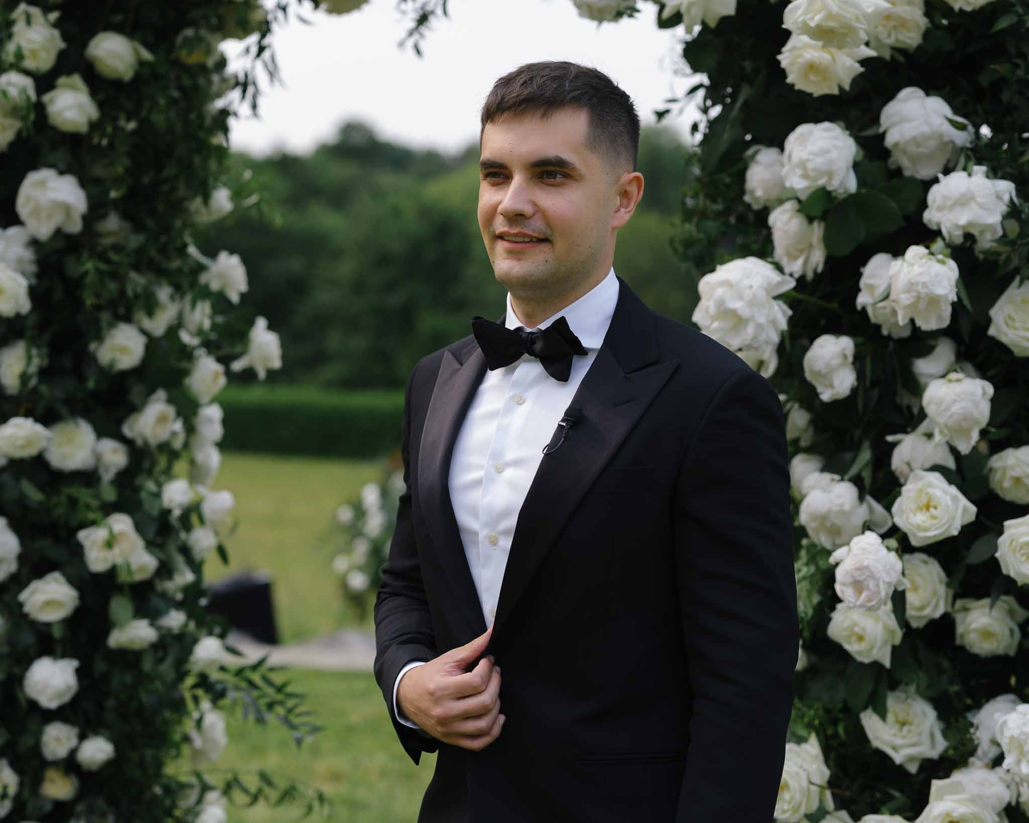 Groom in black tuxedo standing at altar framed by dense white rose and green foliage ceremony arch