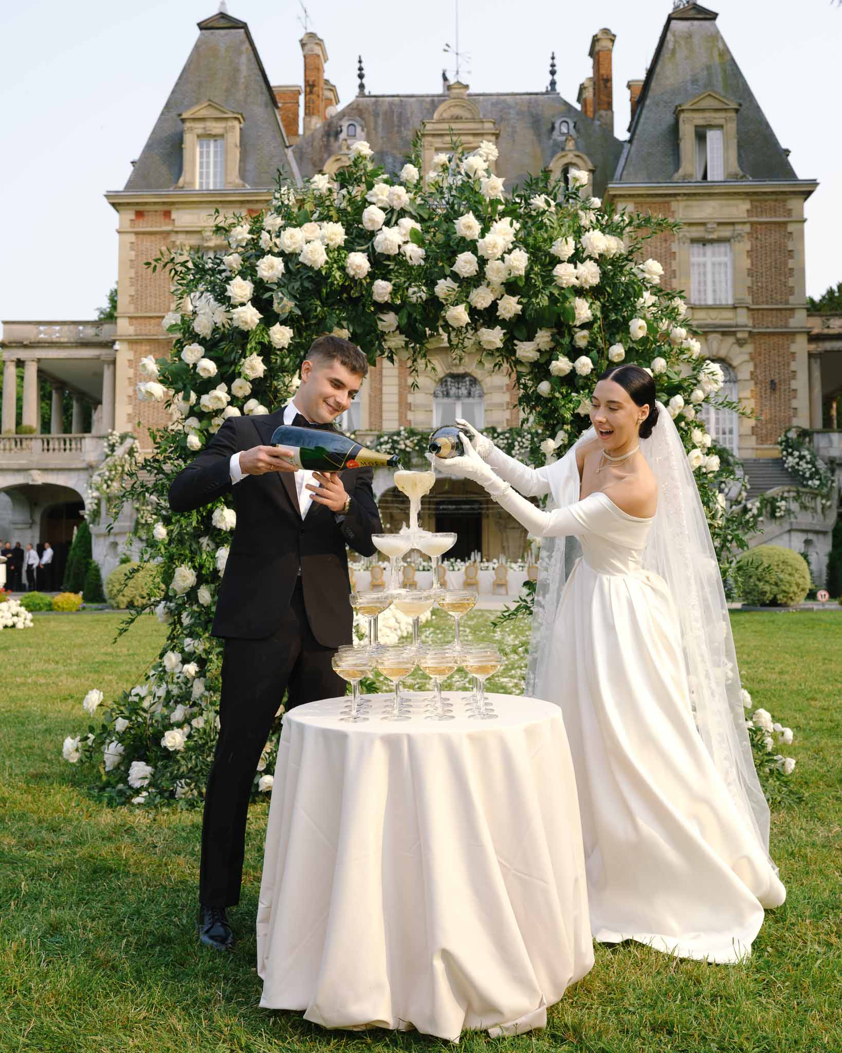 Couple pouring champagne tower before circular white rose arch at chateau facade