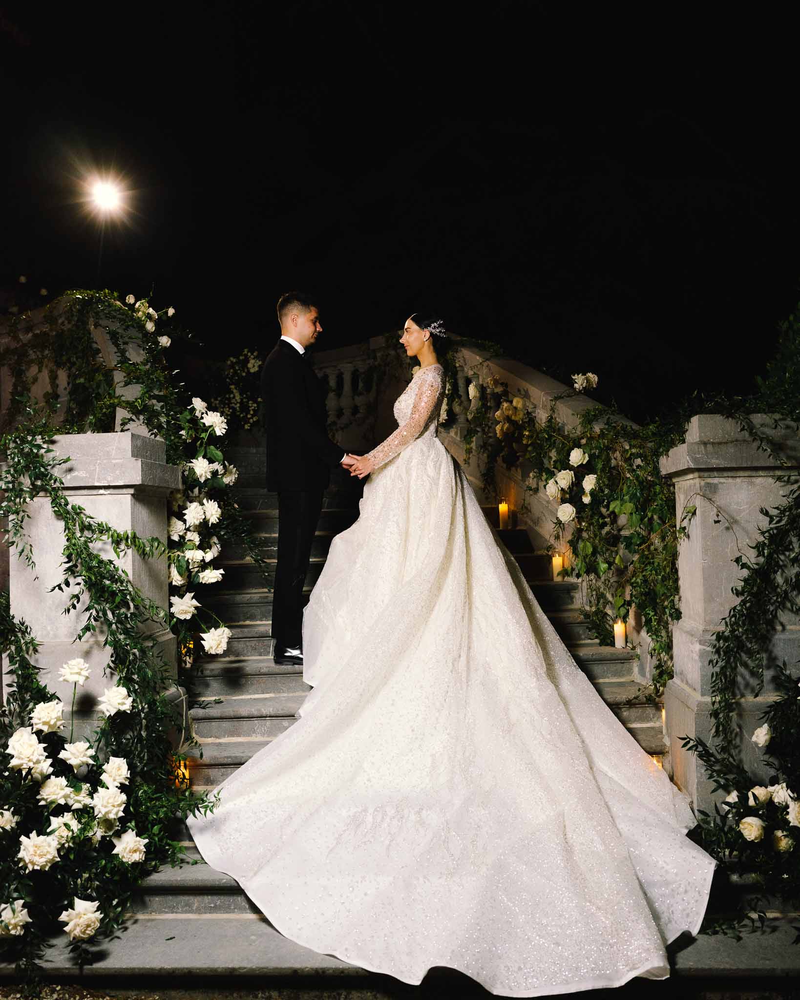 Bride in sparkled gown with cathedral train and groom on candlelit chateau staircase with trailing greenery