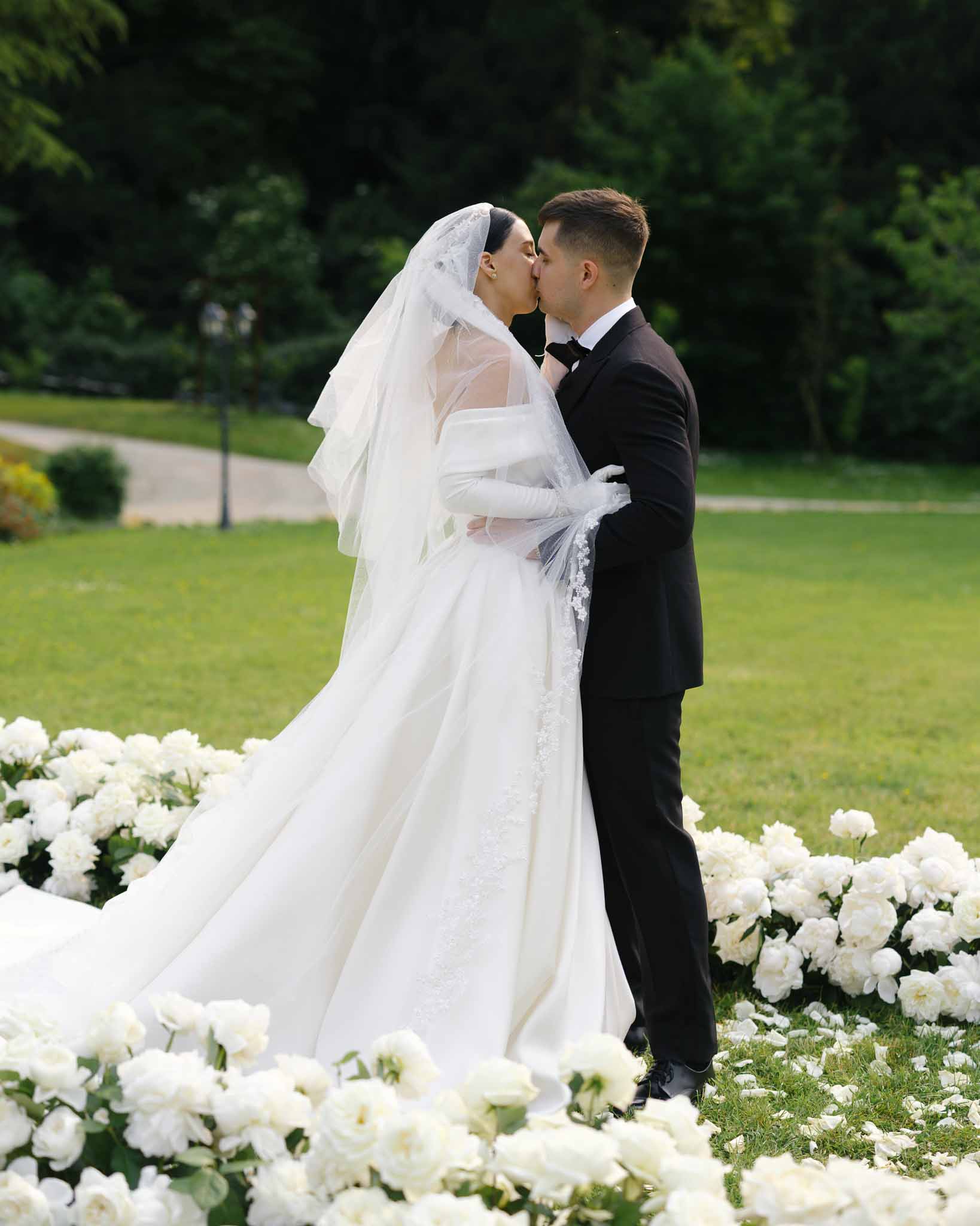 Couple kissing surrounded by white peony and rose ground arc, bride in ivory ballgown with lace gloves and cathedral veil