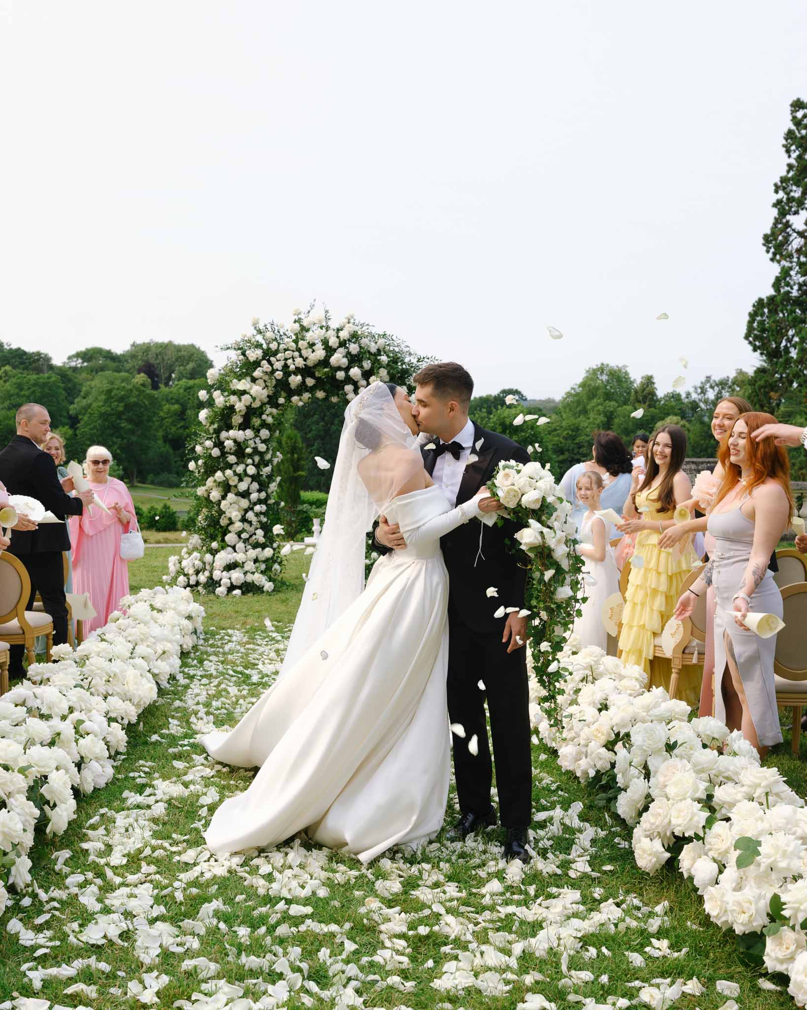 Bride and groom share first kiss under circular white rose arch as guests toss petals on garden aisle