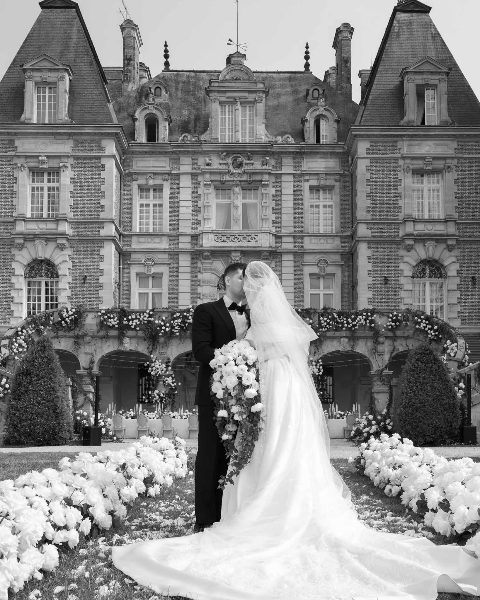 Black and white portrait of bride and groom kissing in front of a baroque chateau with floral-lined lawn