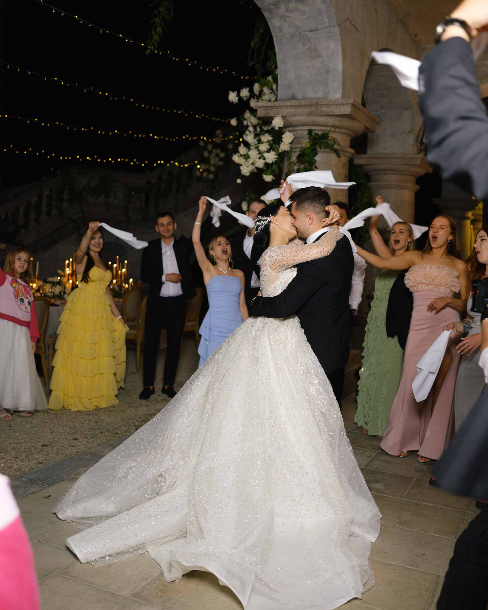 Couple kissing on dance floor as guests wave white handkerchiefs under fairy lights and stone arches