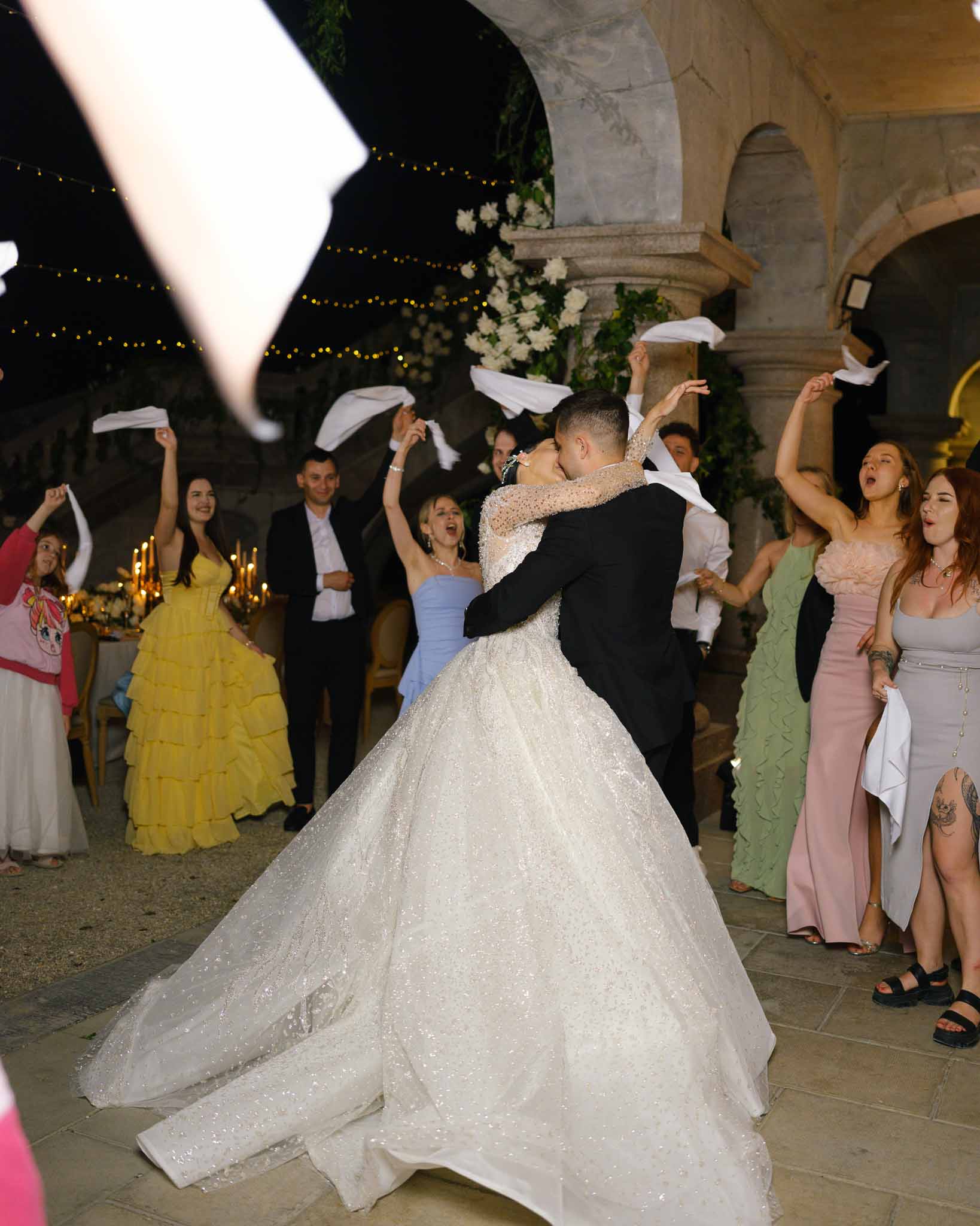 Bride and groom kissing on dance floor as guests wave white napkins under fairy-lit stone arches