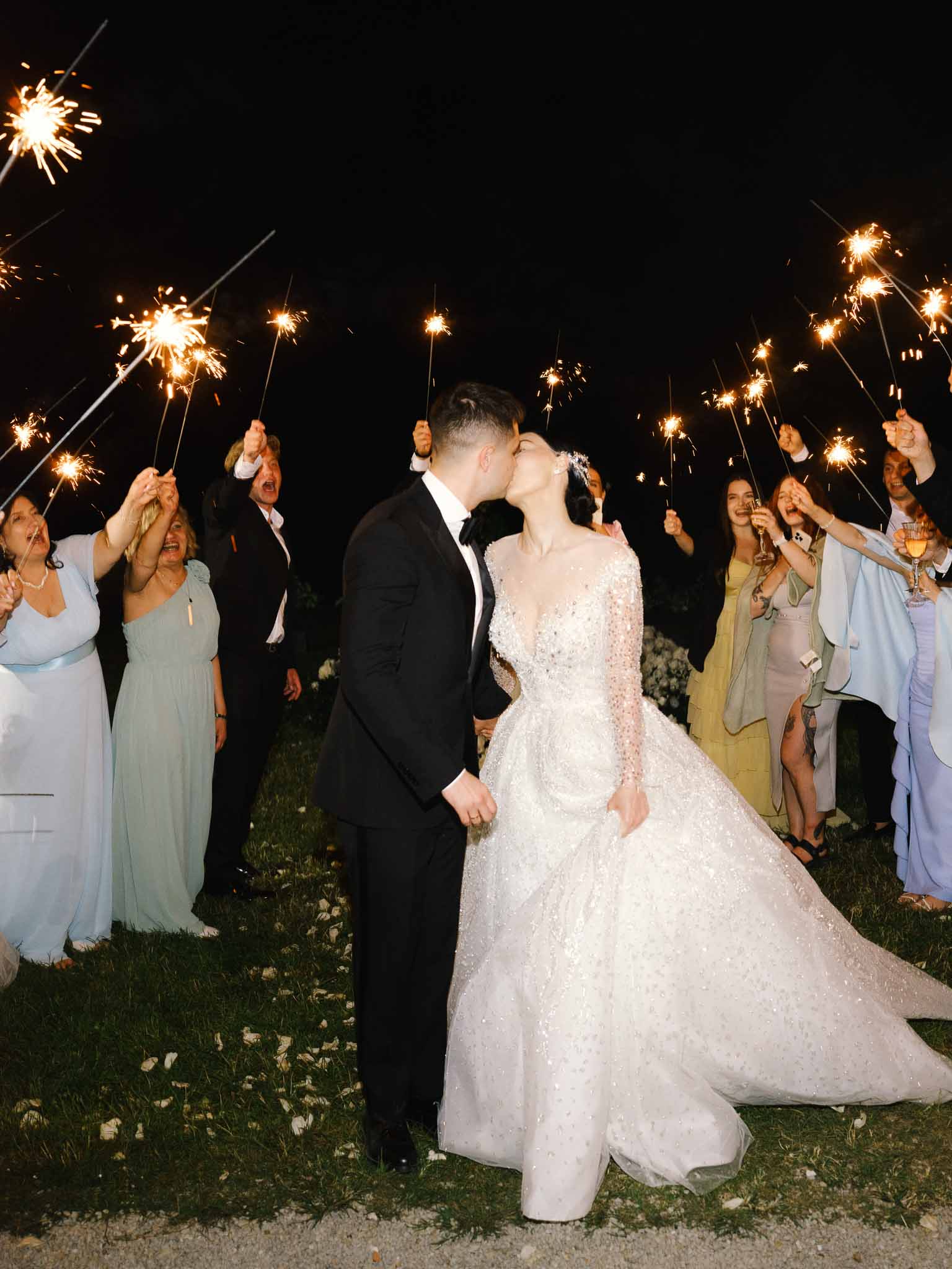 Sparkler send-off corridor with couple kissing, bride in beaded ballgown and groom in tuxedo at night
