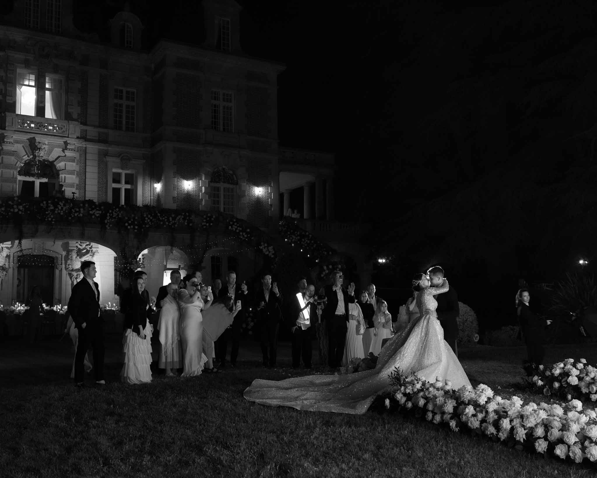 Black-and-white wide shot of bride and groom embracing on chateau lawn at night with guests watching
