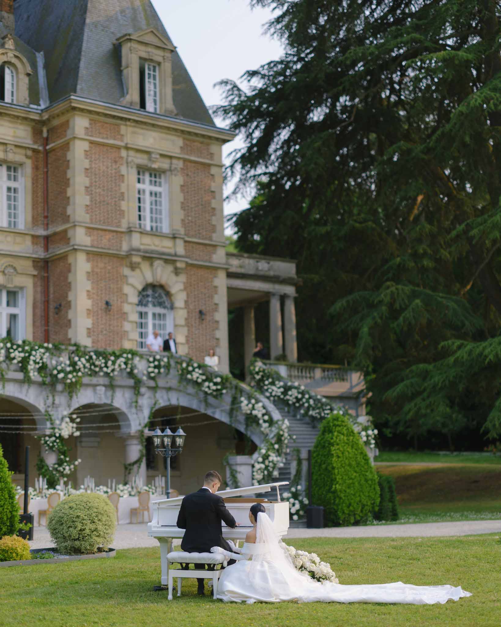 Bride and groom at white grand piano on lawn with rose-draped double staircase behind at chateau