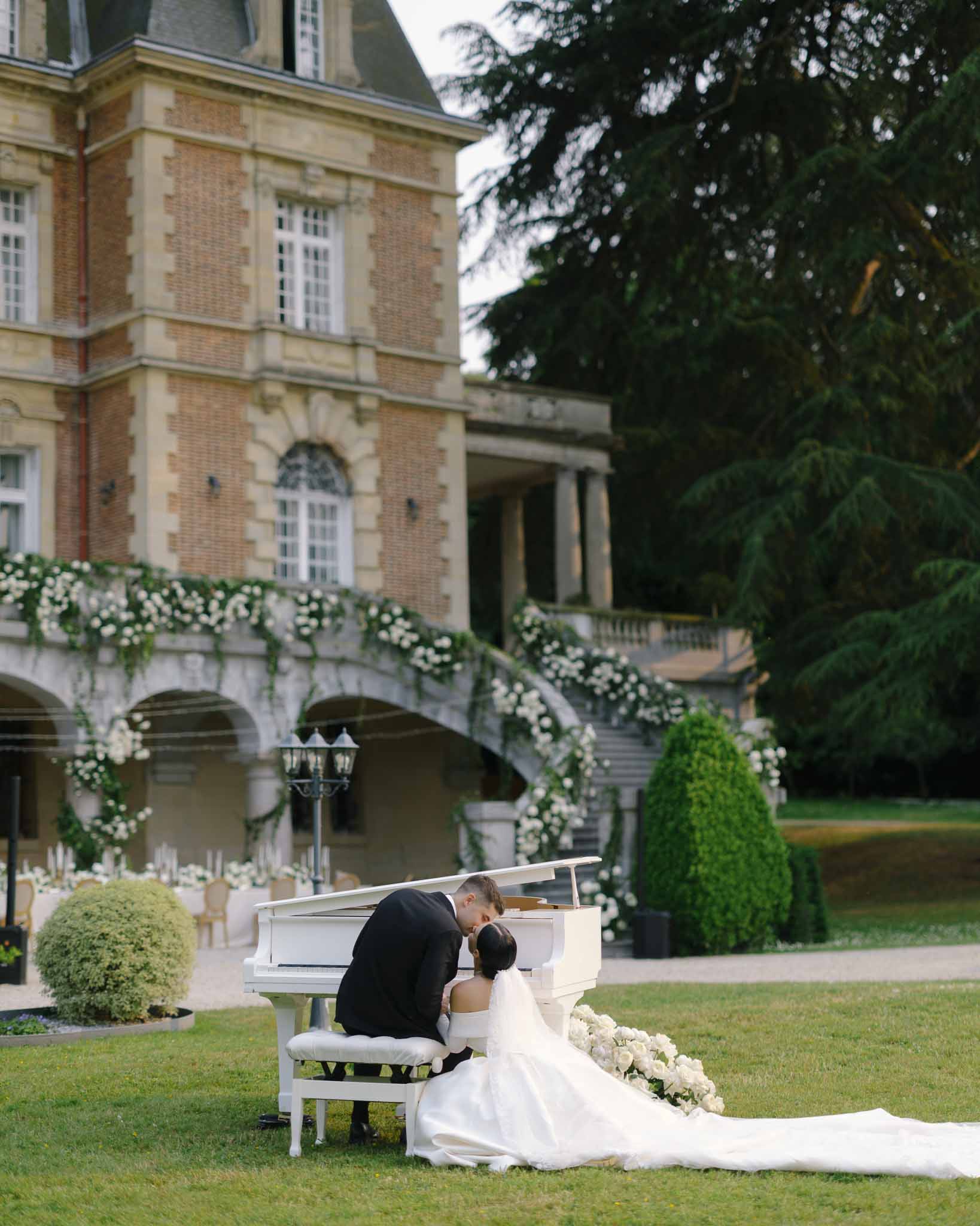 Couple kissing at white baby grand piano on lawn with white rose-draped double staircase at chateau facade