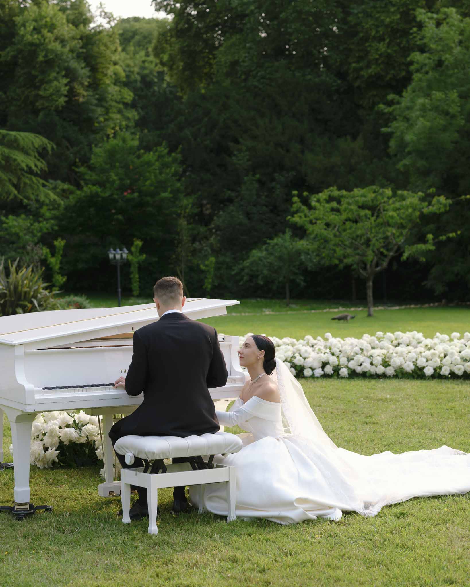 Groom at white grand piano on lawn with bride seated beside in off-shoulder gown with gloves and cathedral veil