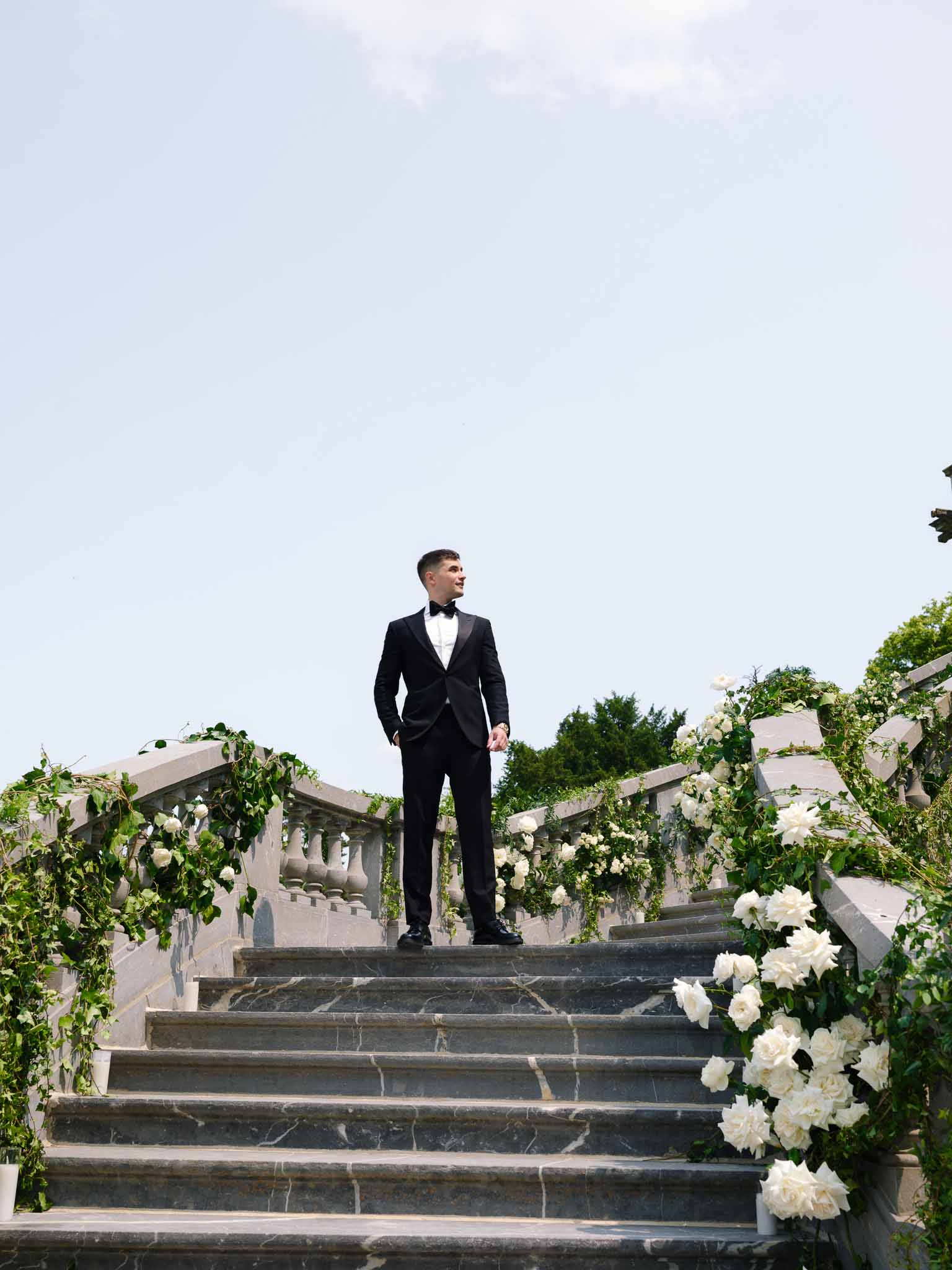 Groom in black tuxedo on marble staircase with trailing white rose and greenery balustrade installations