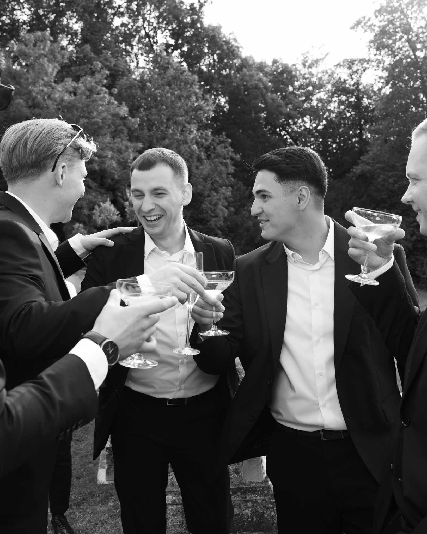 Black and white photo of four groomsmen in dark suits clinking champagne coupes and laughing outdoors
