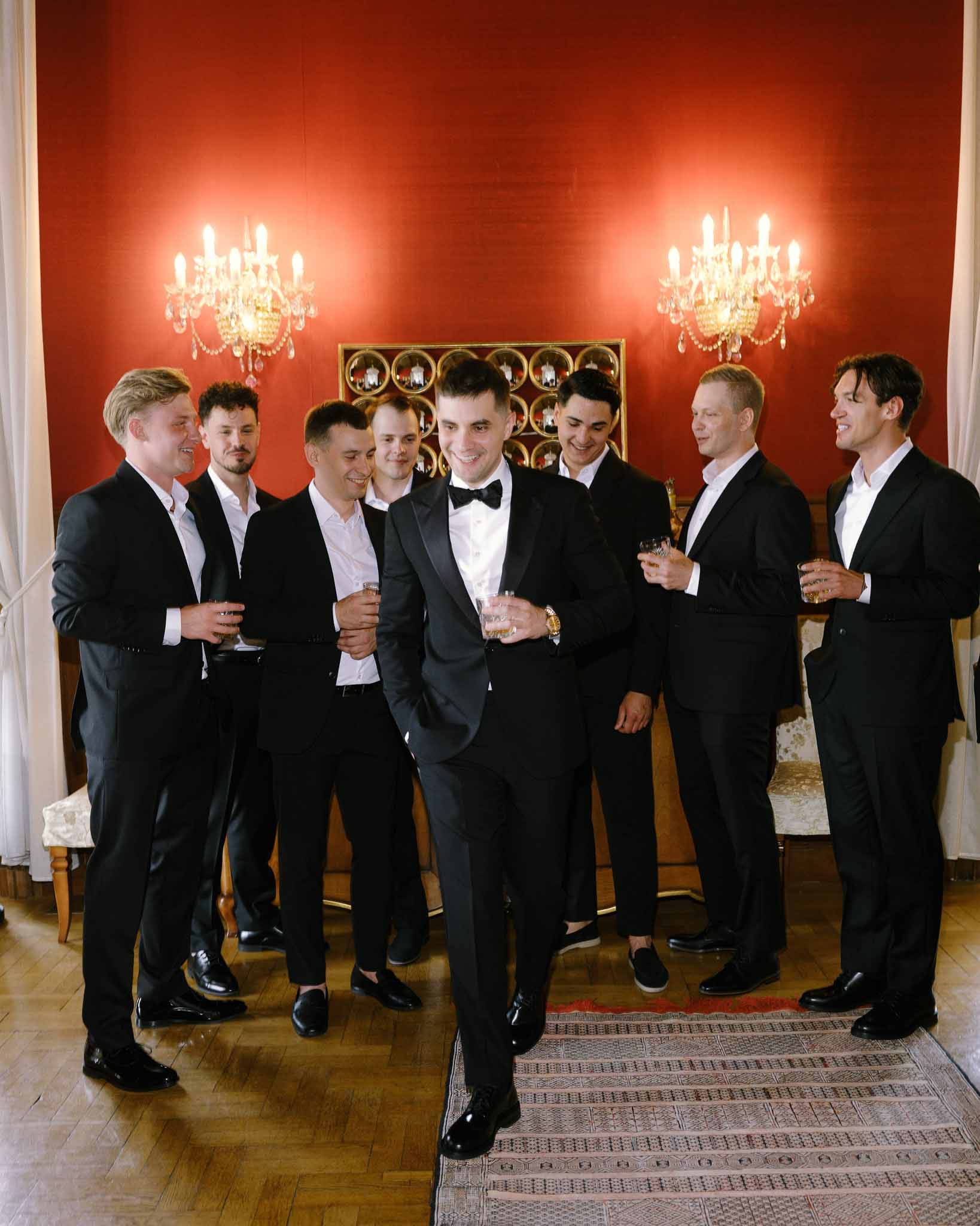 Groom and seven groomsmen in black suits holding whiskey in red-walled room with crystal sconces