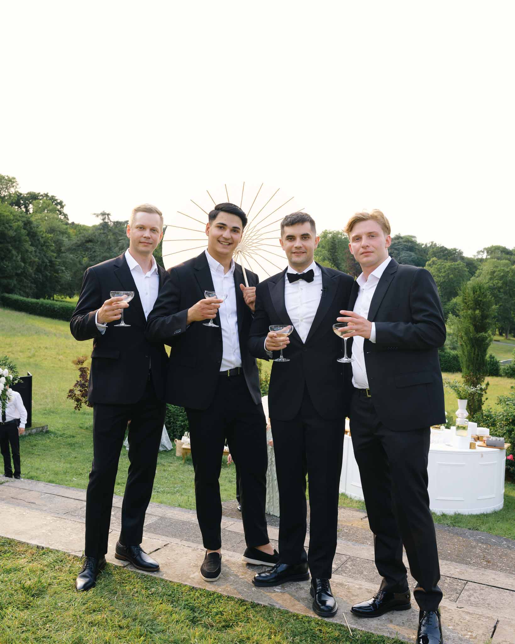 Groom and three groomsmen in black suits holding champagne coupes on stone garden terrace
