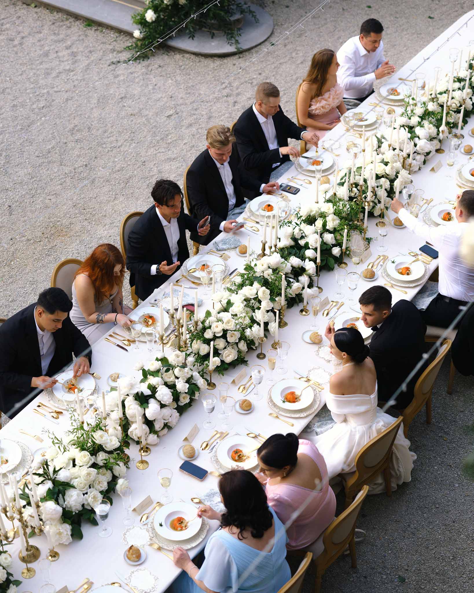 Aerial view of wedding guests seated at long table with white rose runner, gold candelabras, and formal place settings