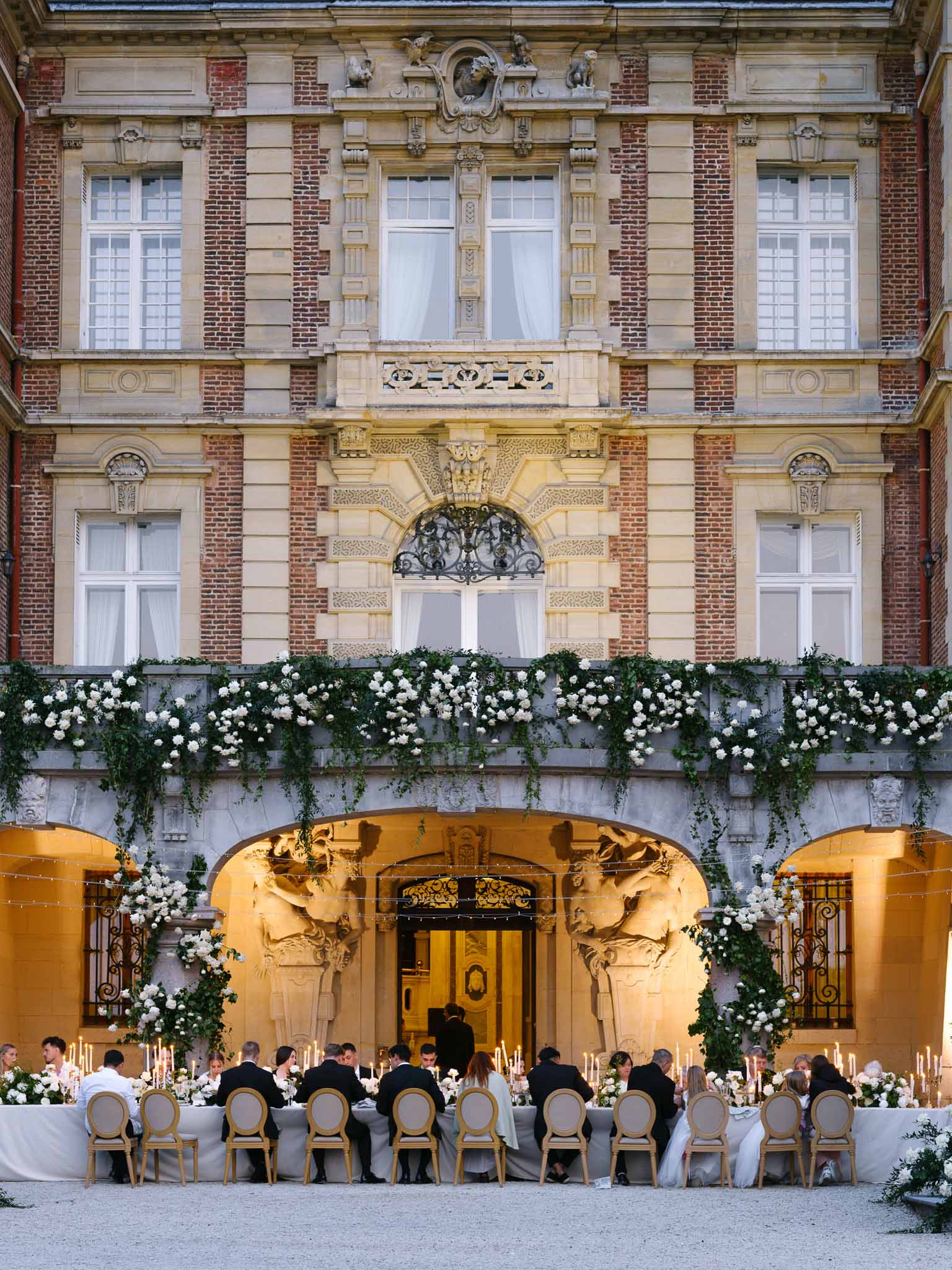 Outdoor wedding reception dinner at a French chateau courtyard with long banquet table, white roses, candles, and formal g...