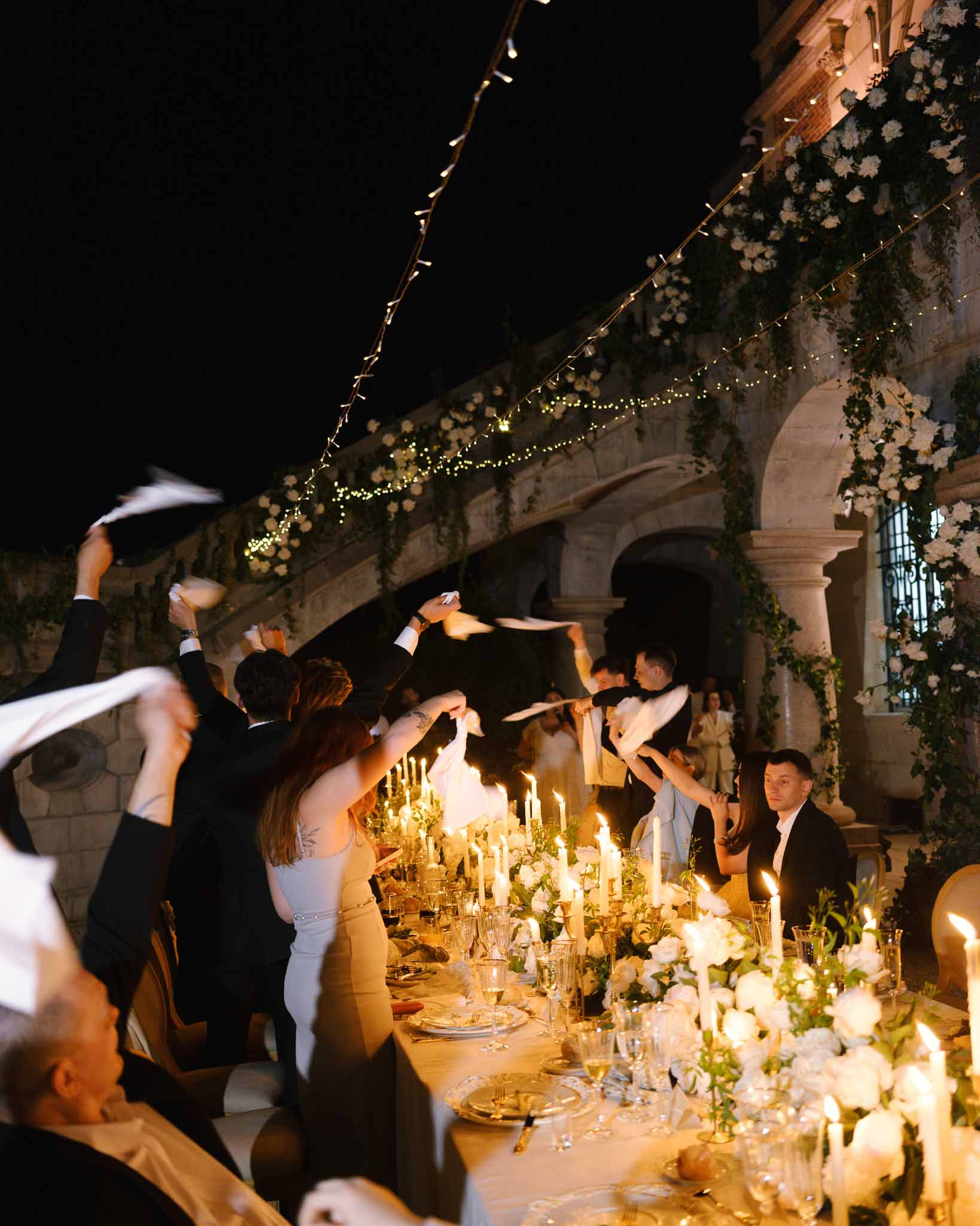 Guests wave white napkins at candlelit table with rose and ivy garland beneath fairy lights at chateau