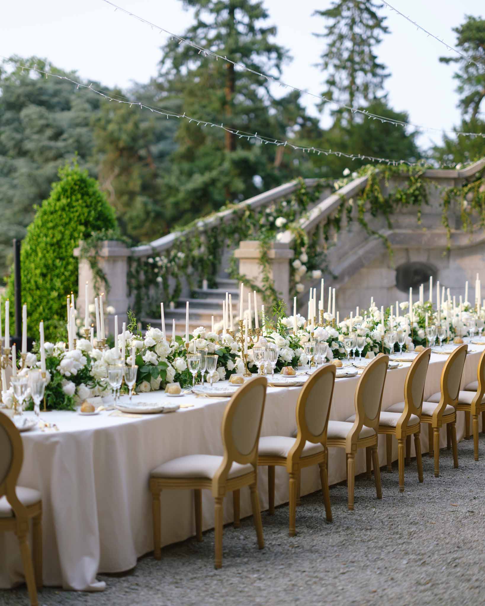 Long banquet table with white roses gold candelabras and Louis XVI chairs before garland staircase
