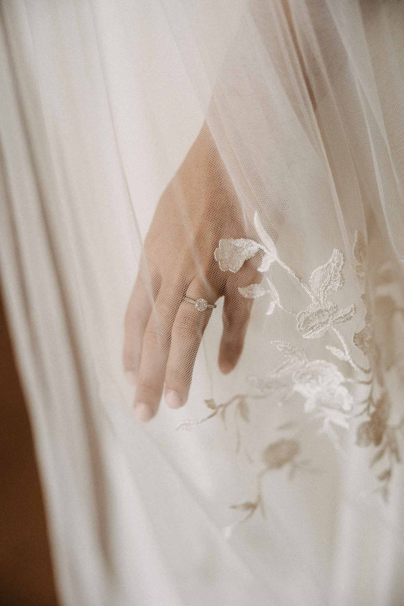 Macro close-up of bride's hand with diamond ring resting on embroidered ivory tulle veil
