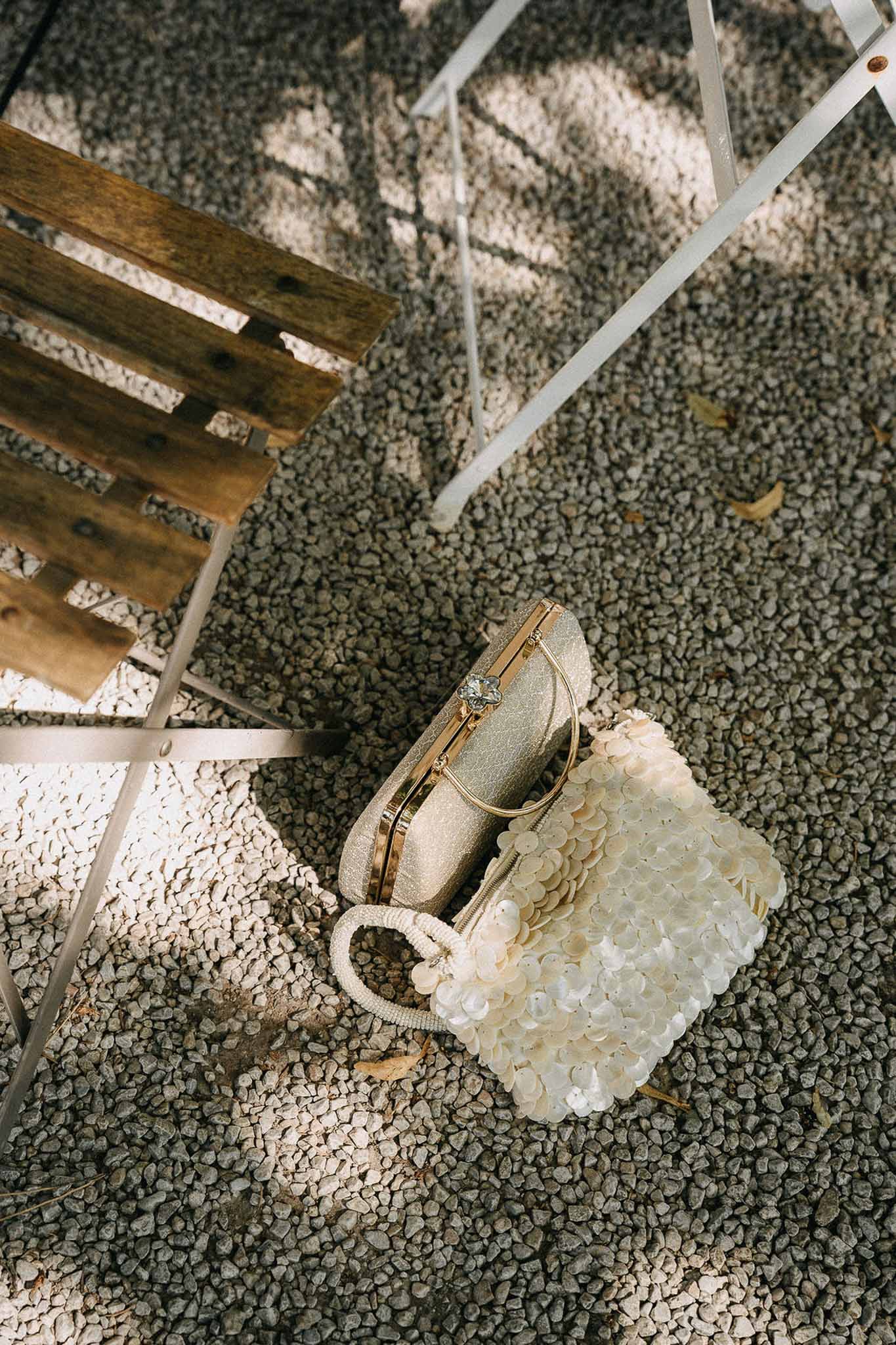 Ivory sequin tote bag and gold glitter clutch styled on gravel beside white bistro chair