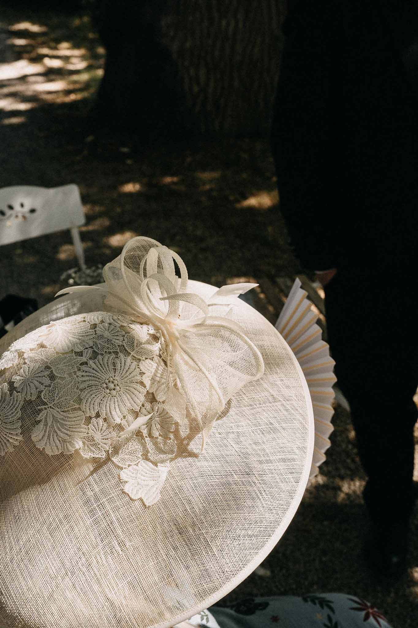 Close-up of cream sinamay fascinator hat with lace applique and tulle bow resting on guest lap at outdoor ceremony