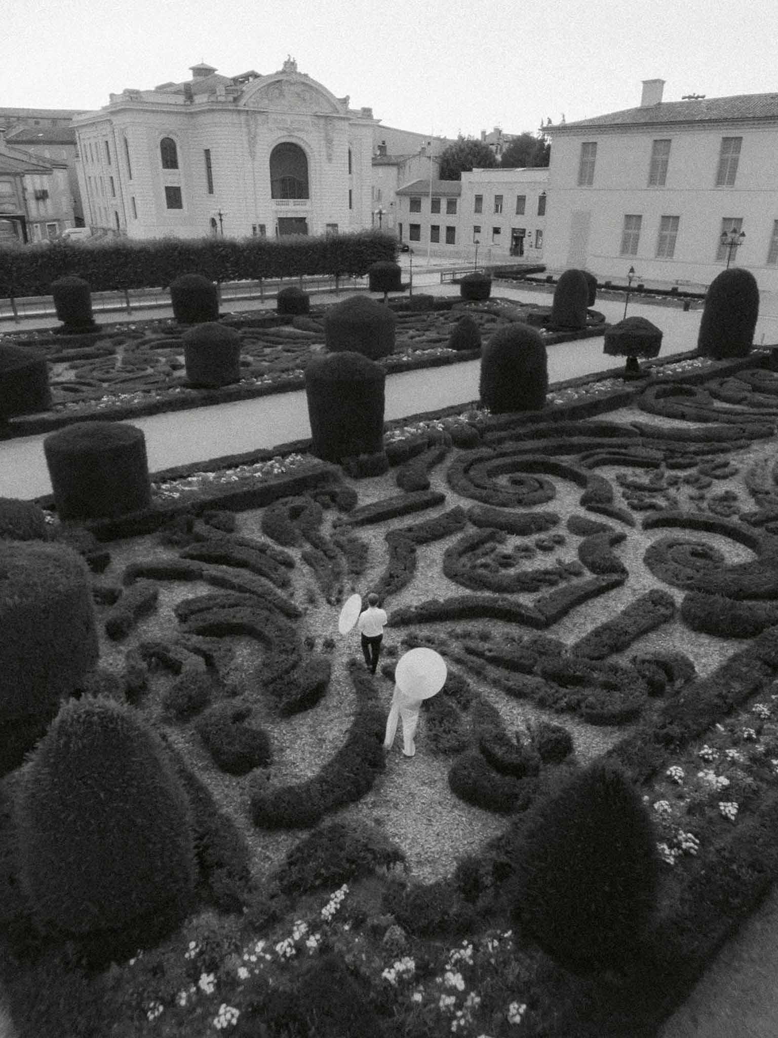 Black and white aerial of couple with white parasols walking through ornamental parterre garden at chateau