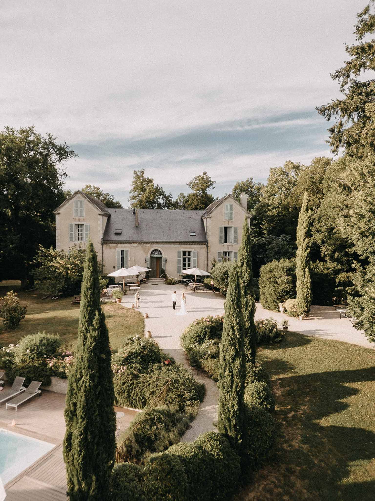 Aerial view of stone manor with blue-grey shutters, cypress trees, formal landscaping, and pool