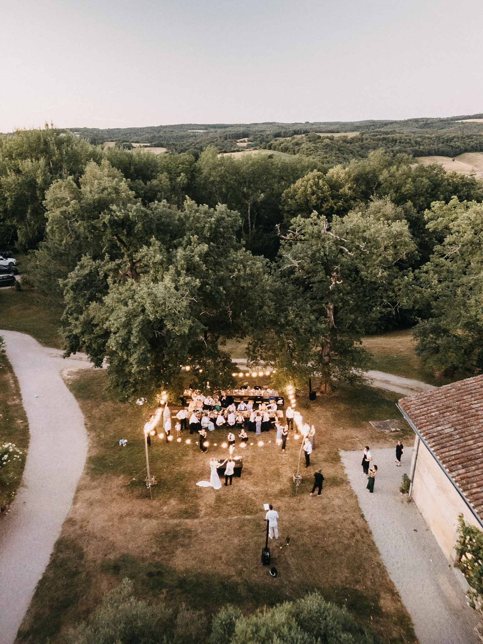Aerial dusk view of couple dancing on lawn beside U-shaped reception tables under festoon lights