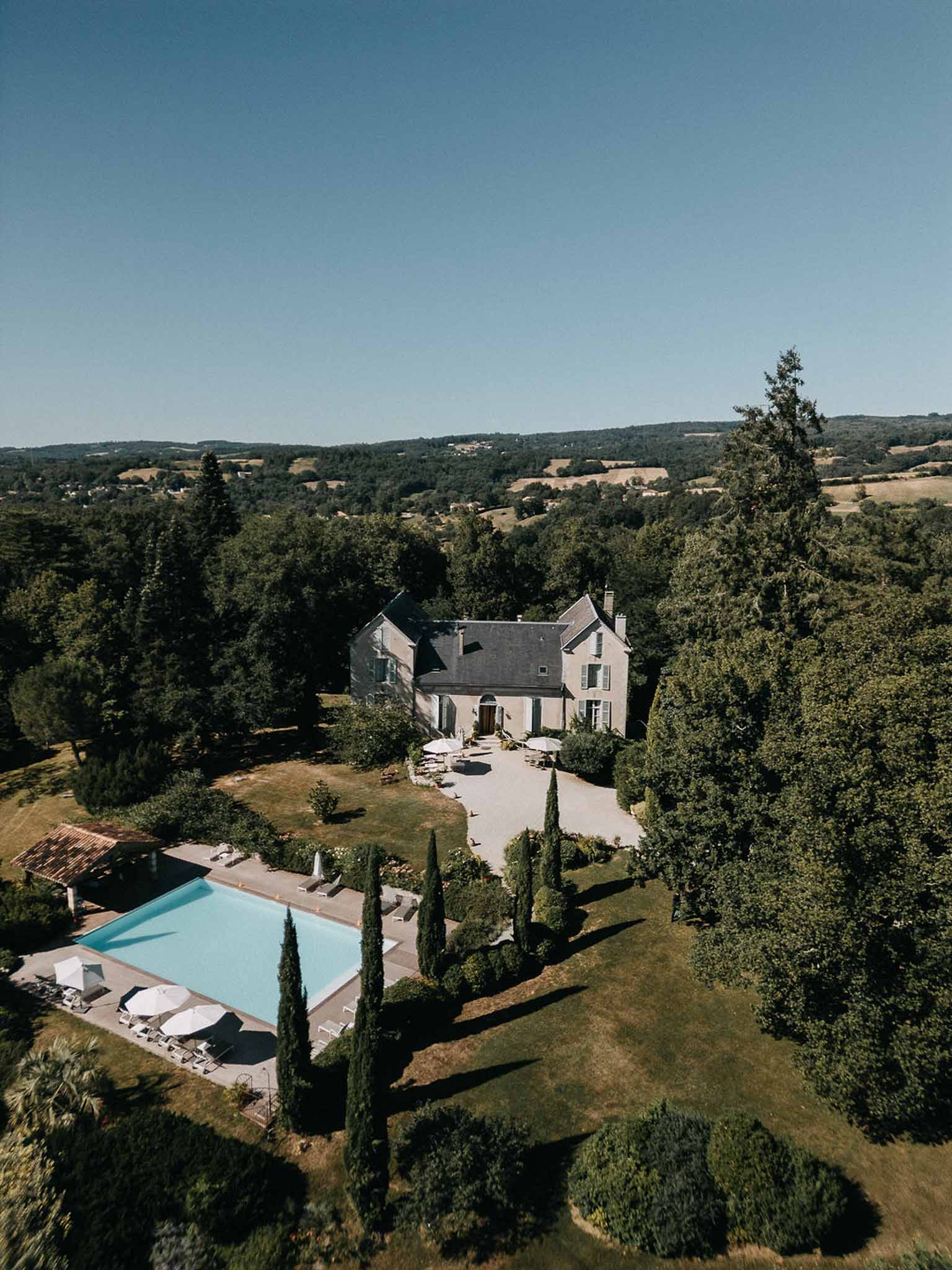 Aerial drone view of French stone manor with swimming pool, cypress trees, and rolling countryside