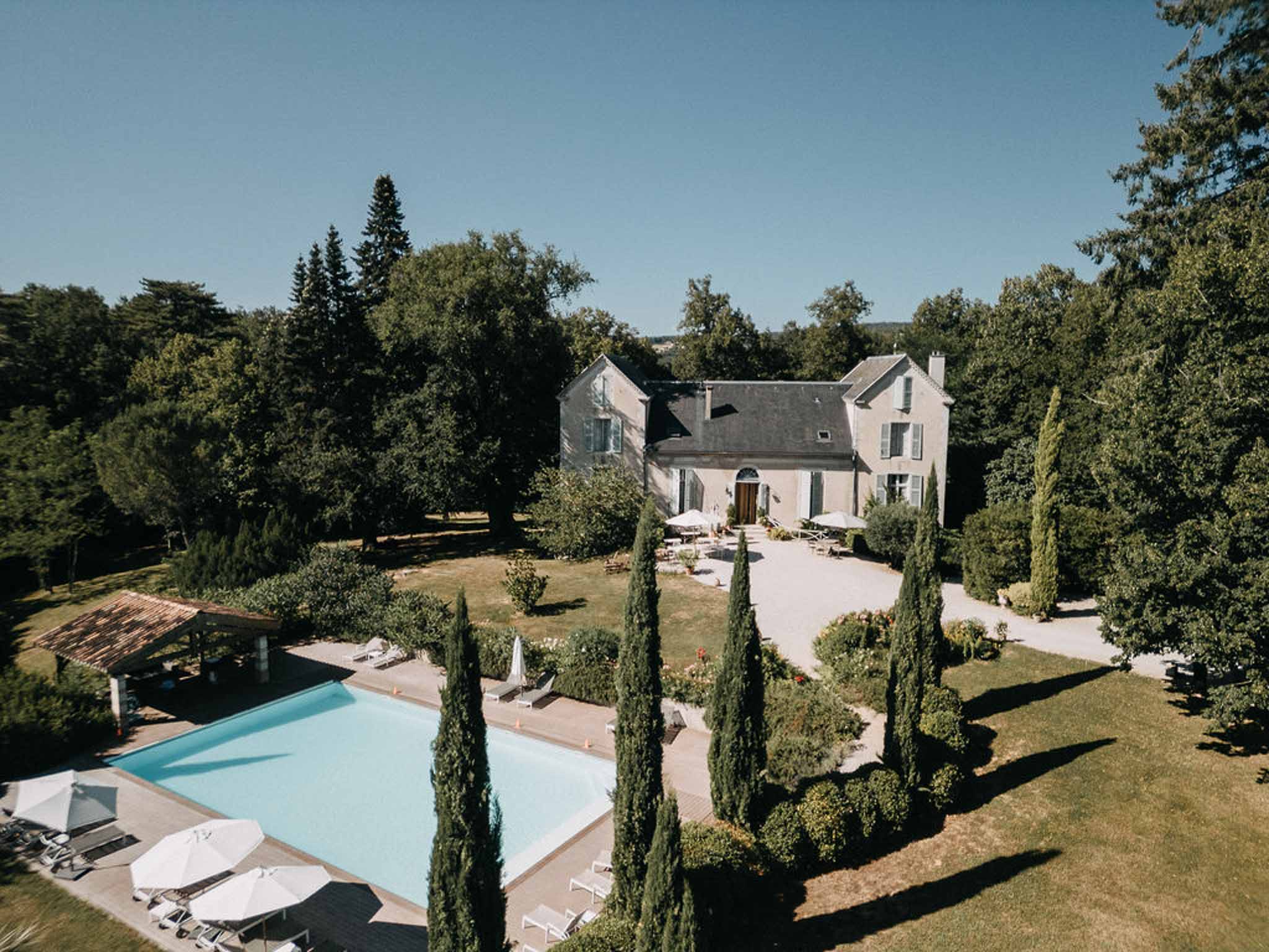 Aerial view of manor with slate roof, pool with white loungers, and cypress-lined pathway to entrance