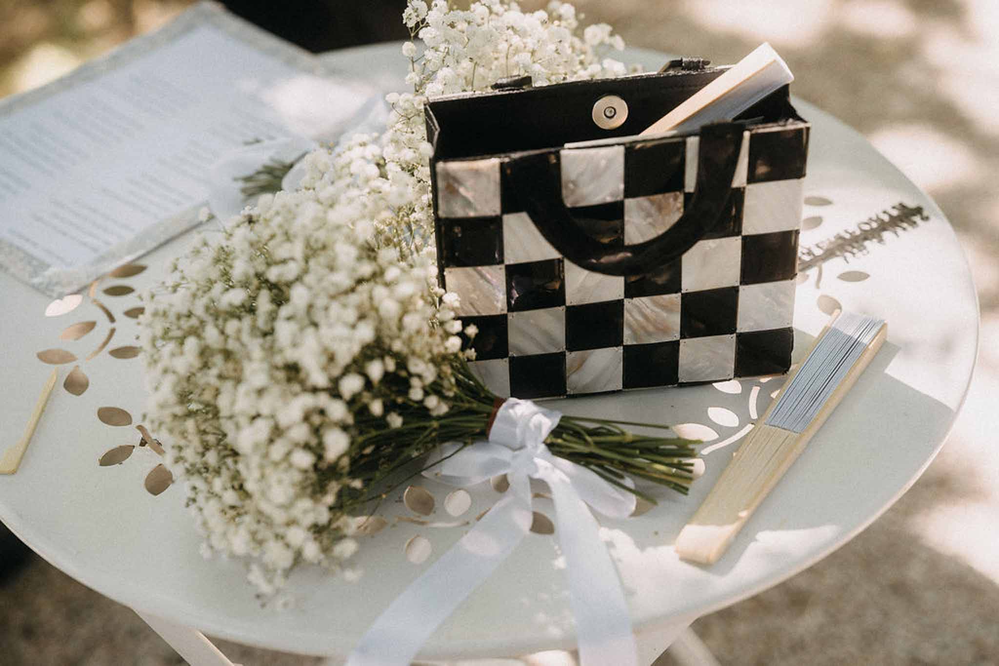 Bridal accessories on garden table with baby's breath bouquet, checkered clutch bag, and wooden hand fan